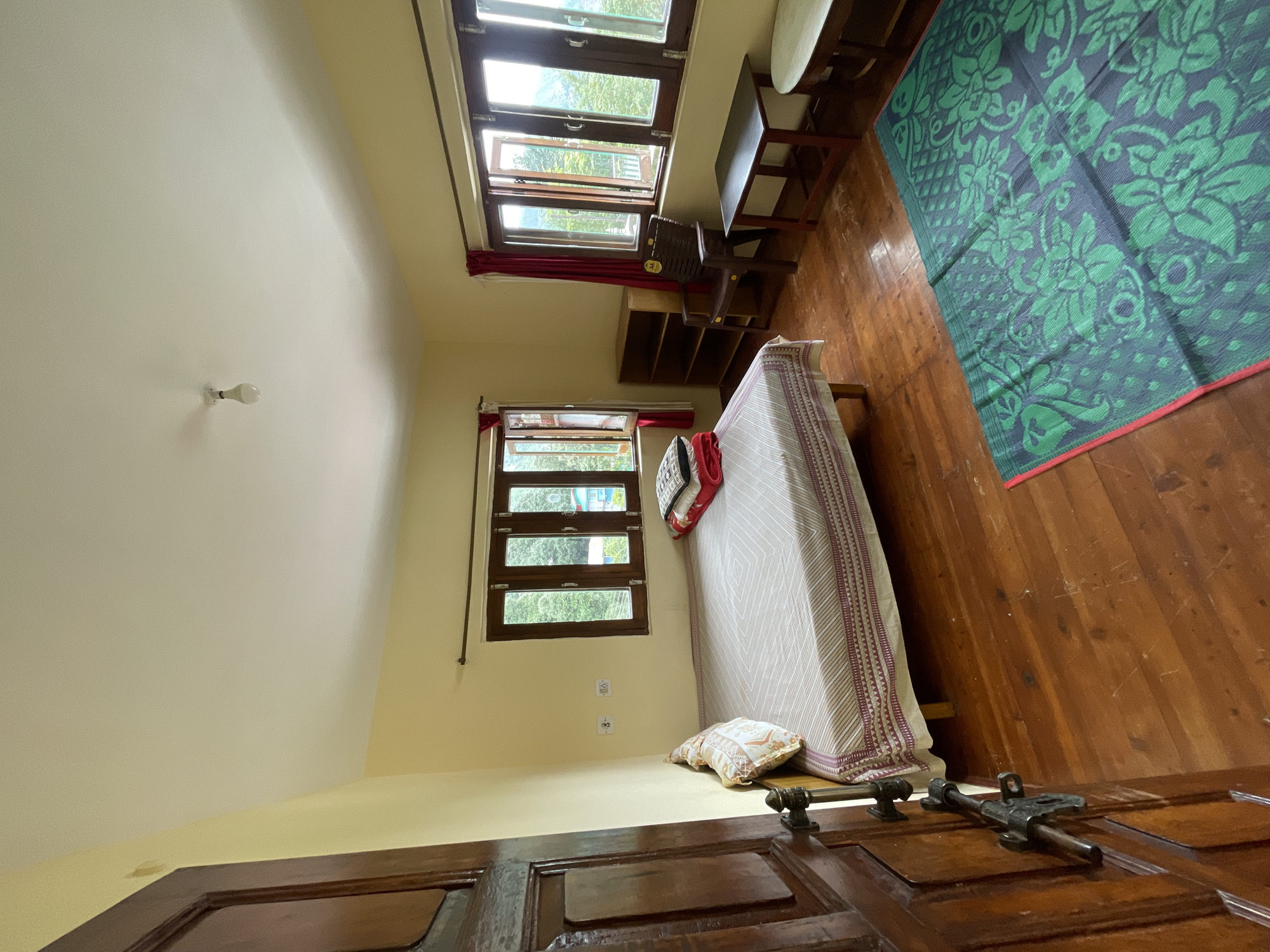 A brightly lit, modestly furnished bedroom in Bhagsu, India, is captured in this photograph. The room features a wooden floor and pale yellow walls. On the left side of the frame, a dark wooden door with ornate metal hardware stands ajar, revealing a glimpse of the hallway beyond.
Dominating the center of the room is a single bed with a wooden frame, covered by a light-colored, patterned bedsheet. A few pillows and folded blankets are neatly arranged on the bed. Beside the bed, a single pillow with an intricate pattern rests against the headboard.
To the right of the bed, a large wooden-framed window offers a view of lush green trees and a distant mountainous landscape. The window has multiple panes and is partially open, allowing natural light to flood the room. Red curtains are drawn on either side of this window.
Further to the right, a dark brown plastic chair sits next to a narrow, dark wooden shelf unit with multiple compartments. In front of this area, a dark wooden table with a dark top stands adjacent to a small, round, light-colored side table.
In the foreground, a large green mat with a floral pattern is laid out on the wooden floor. The overall impression is of a clean, simple, and functional bedroom, likely a guest room in a guesthouse or homestay in the scenic town of Bhagsu. The ceiling is white with a single, exposed lightbulb. Two electrical outlets are visible on the wall between the door and the window.