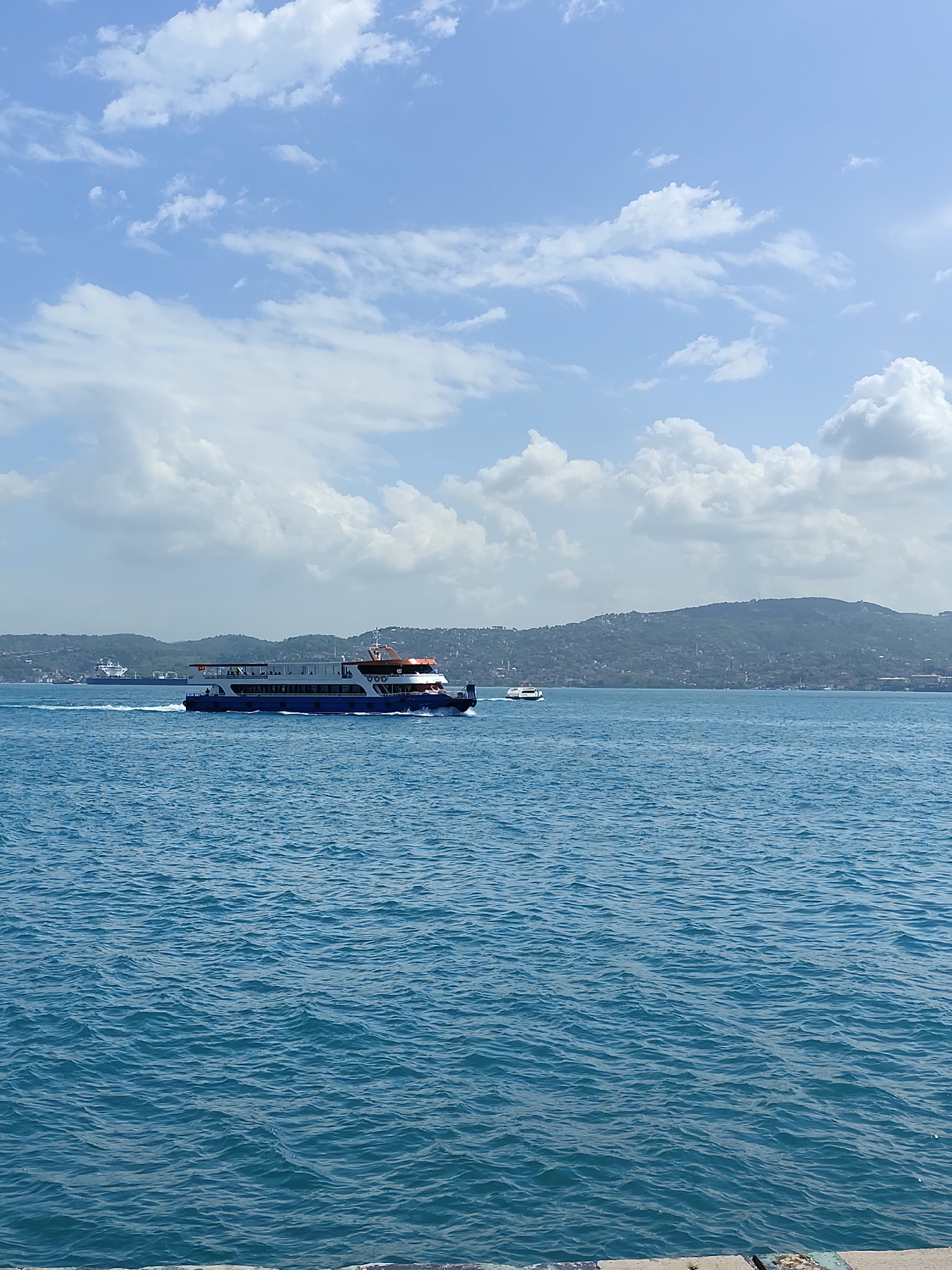 The image depicts a seascape, likely taken on a clear day with a predominantly blue sky adorned with scattered cumulus clouds. The sea, a deep blue hue, dominates the lower portion of the frame. On the water, a passenger ferry, dark blue and white with orange accents, is moving towards the right side of the image. A smaller vessel is also visible in the distance, trailing behind the ferry.
In the background, a hilly landscape is visible, likely a city scape on a distant shore. The specific location is unknown.
The foreground appears to be the edge of a pier or dock, evidenced by a visible concrete border. The overall scene is calm and serene, capturing a typical day on the water.