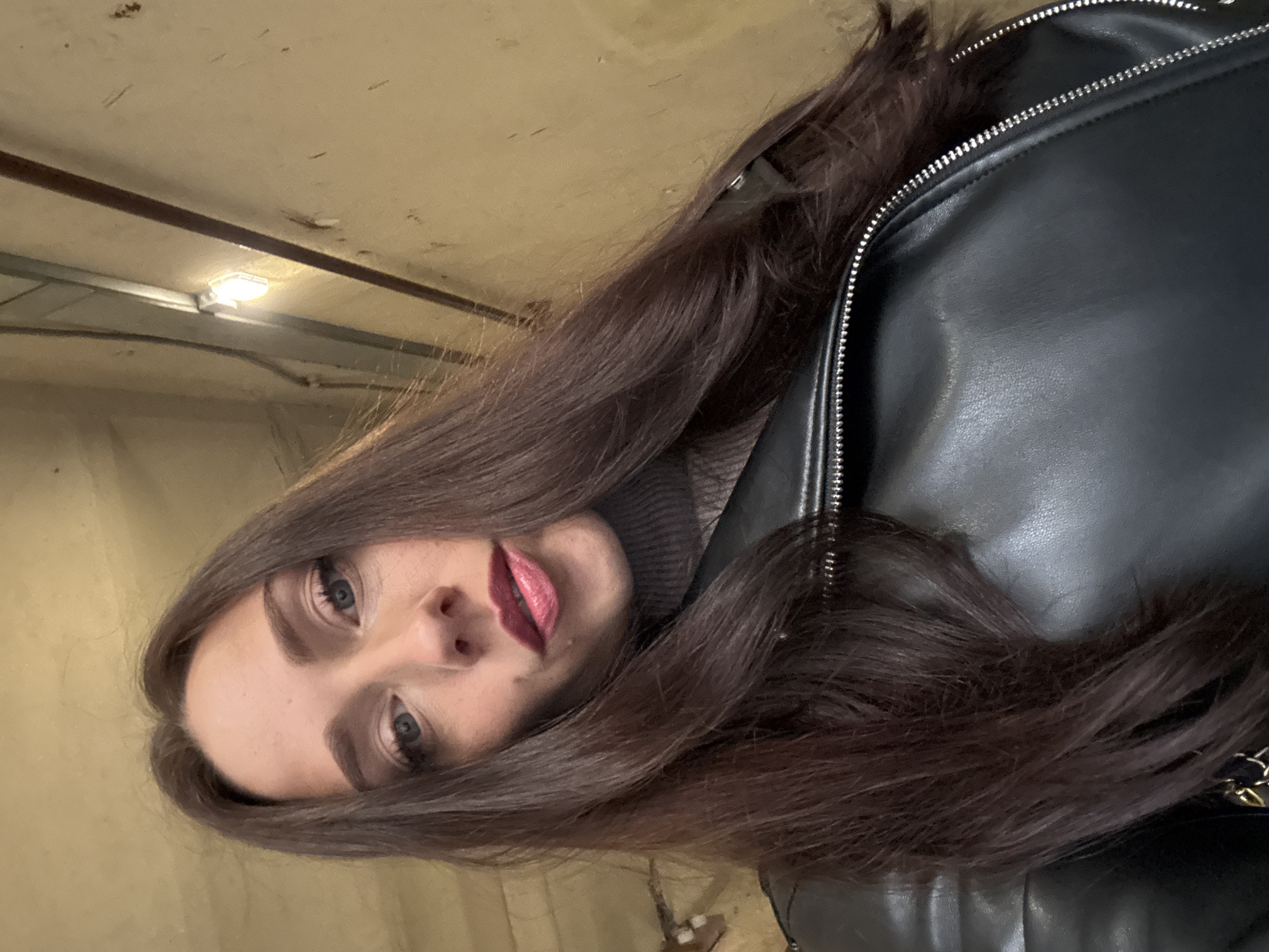 A woman with long, dark brown hair is looking up at the camera. She is wearing a gray turtleneck sweater and a black leather jacket with a silver zipper. Her lips are painted with a reddish-pink lipstick. The background appears to be an interior hallway with a yellow-tan wall and a light fixture on the ceiling. The scene is likely in St. Petersburg, Russia, based on the provided metadata.