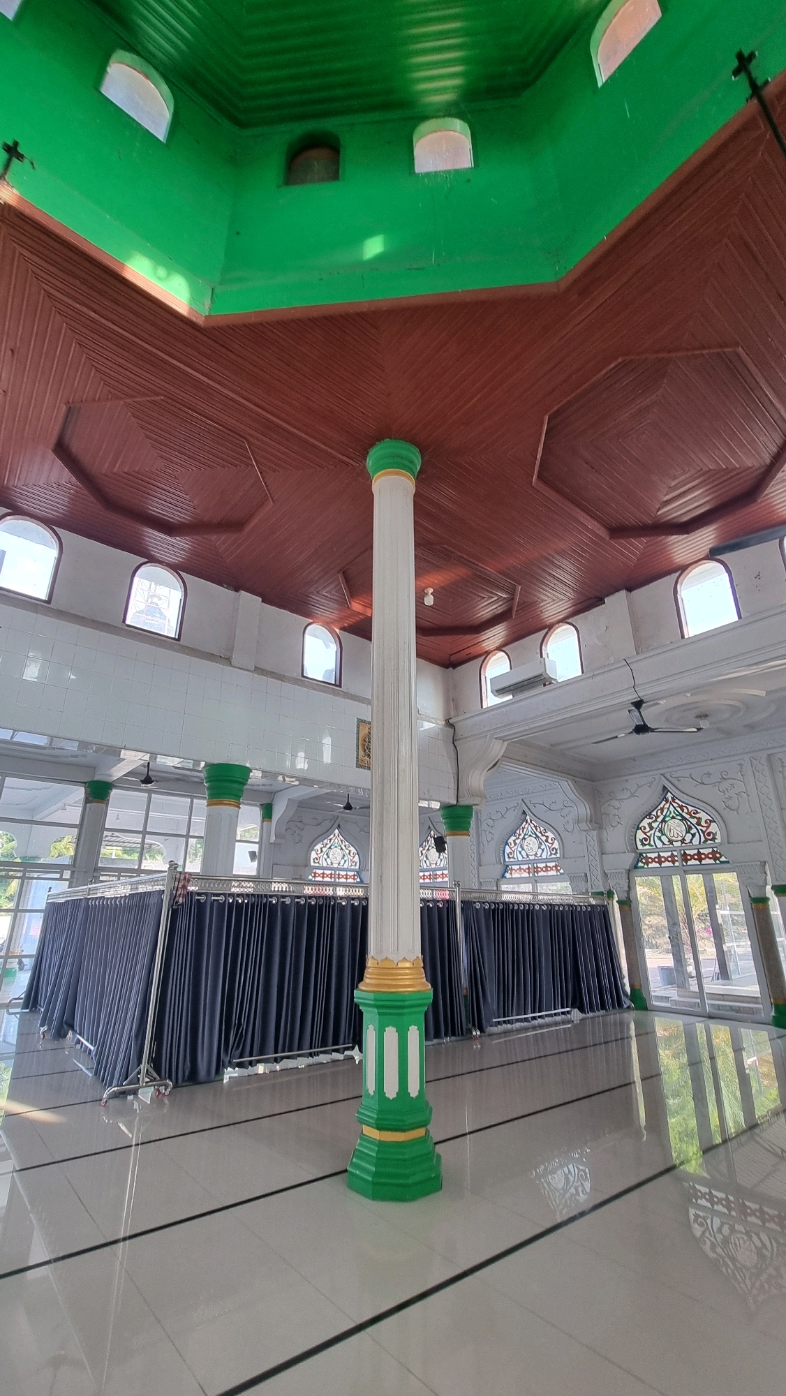 This image depicts the interior of a building with a green ceiling and a brown wooden paneled ceiling. A large white pillar with green and gold accents stands in the foreground, supporting the ceiling. The walls are white with arched windows. In the background, dark blue curtains are drawn, concealing an area. The floor is tiled with a glossy finish and has thin black lines. The scene is well-lit, with natural light entering through the windows, creating reflections on the floor. The architectural style suggests a place of worship or a significant communal building, possibly in Blangrambong, Indonesia, given the context. There are no people visible in the image.