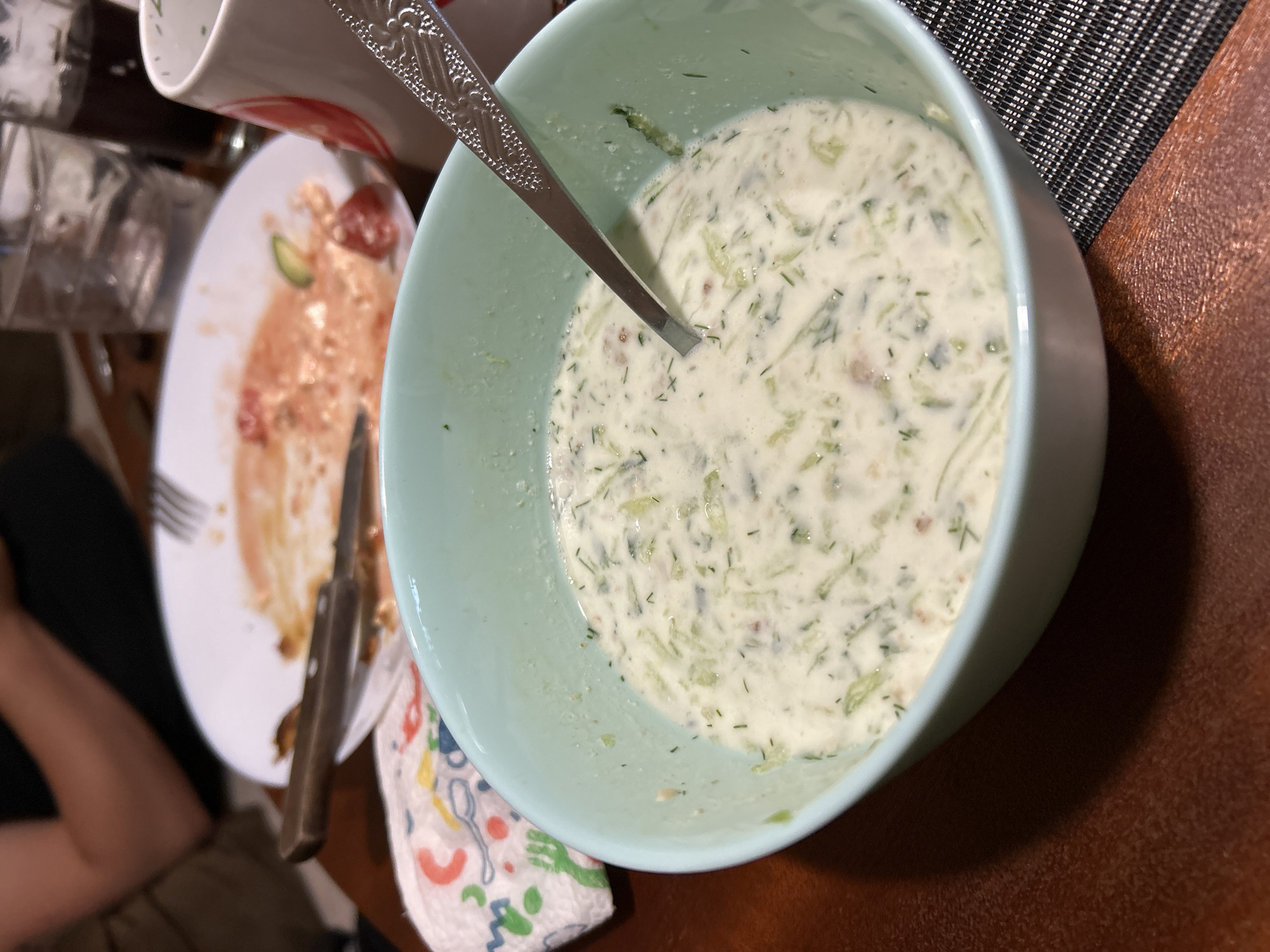 The image shows a close-up of a light blue bowl filled with a creamy white soup containing chopped green herbs and what appear to be cucumber pieces. A silver spoon rests in the bowl. In the background, a dirty white plate with remnants of food, a knife, and a napkin with colorful cartoon drawings are visible. Further back, there are two glasses, one partially filled with a clear liquid and another with a dark liquid. A white mug with a red graphic is also present. The setting appears to be a casual dining environment, possibly a home or restaurant, on a wooden table. There is no specific indication of the city of Sofia, Bulgaria.