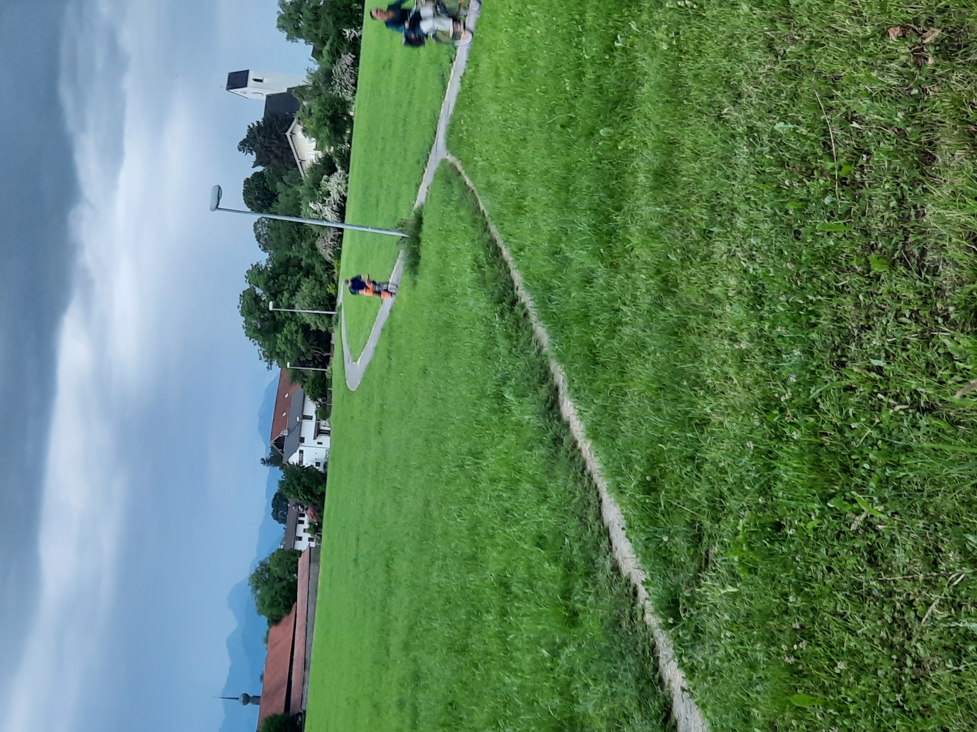 The image depicts a pastoral scene in Rosenheim, Germany, with a large expanse of vibrant green grass dominating the foreground and midground. A winding paved path snakes through the grass, leading the viewer's eye towards a cluster of buildings and trees in the background. The sky is overcast with dramatic, dark clouds, suggesting impending rain or a storm.
In the midground, two individuals are on bicycles, navigating the path. One cyclist, wearing a blue shirt, is further away, moving away from the viewer. The second cyclist, closer to the foreground and on the right side of the path, is partially visible and appears to be wearing a dark top and white pants, riding towards the viewer.
Behind the cyclists, a scattering of trees and white-flowered bushes are visible. A white church with a black steeple stands prominently among the trees, its architecture suggesting a historic building. To the left of the church, other buildings with red-tiled roofs and white facades are discernible, appearing to be part of a village or small town. In the far distance, a range of hazy blue mountains forms the horizon. The overall atmosphere is serene, despite the ominous sky, conveying a sense of rural tranquility.