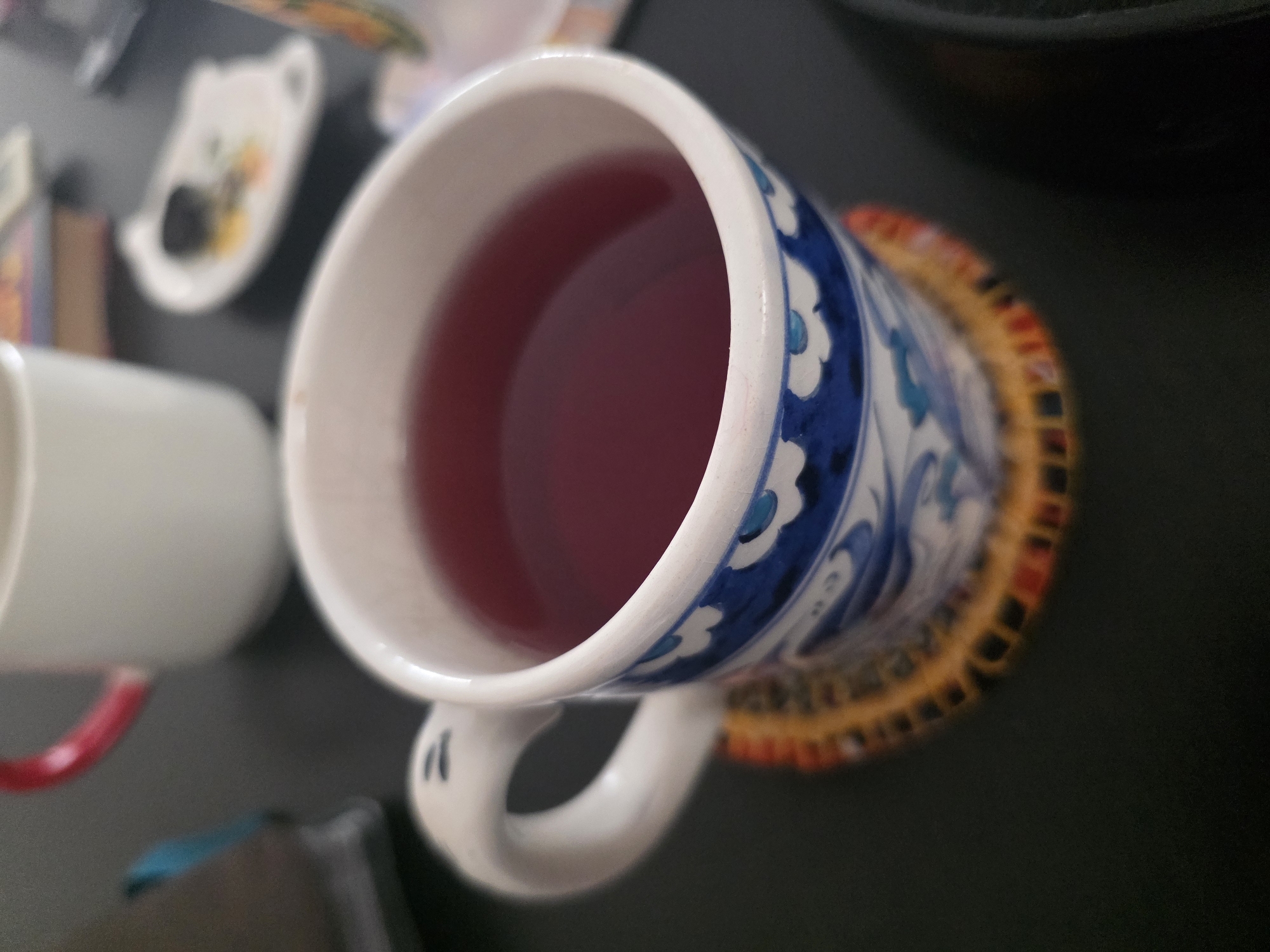 The image shows a close-up of a mug filled with a dark red beverage, likely tea or juice. The mug has a white exterior with blue and white floral patterns around the base and on the handle, which is also white. The mug sits on a coaster with an orange and yellow woven pattern. In the background, out of focus, is another white mug with a red handle. To the right of the mug, there is a small, white, leaf-shaped dish with some dark object in it. The overall setting appears to be a tabletop, suggesting an indoor environment. There is no visible indication of the specific location in Bishkek City, Kyrgyzstan.
