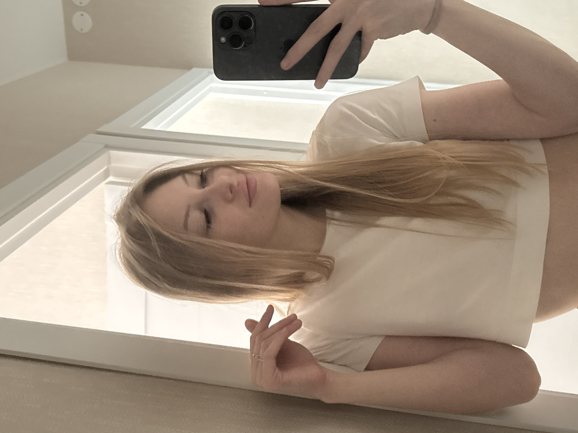A young woman with long blonde hair is taking a mirror selfie in a room that appears to be a bathroom or dressing area. She is wearing a cream-colored crop top and has a ring on her left hand. Her right hand is holding a dark-colored smartphone, possibly an iPhone, which is angled to capture her reflection. The background shows a door frame, a wall with a textured finish, and part of another doorway or window. The lighting suggests an indoor environment with natural light coming from the right. There are no discernible indicators of the city of Perm, Russia in the image.