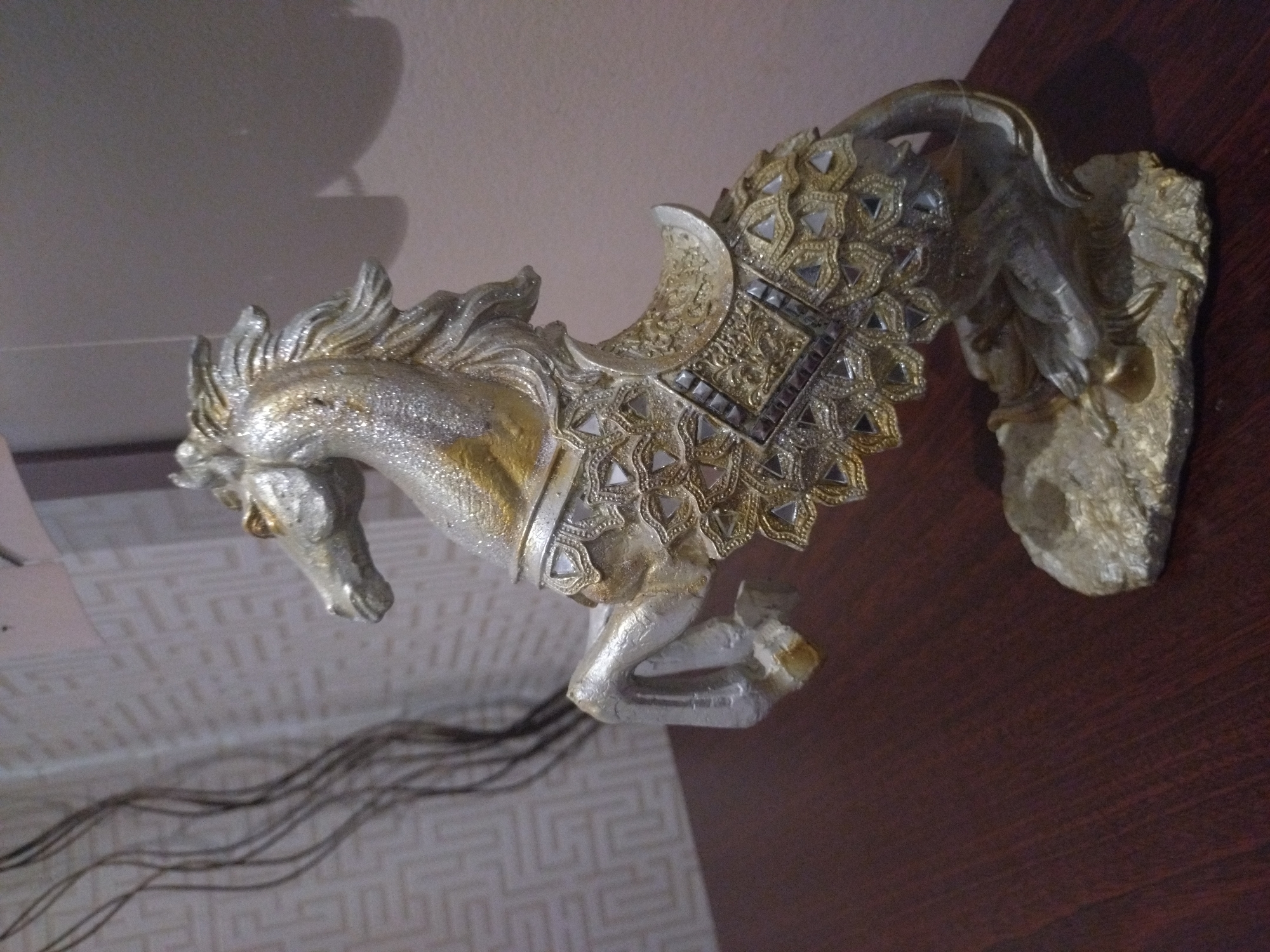 A detailed photograph captures a metallic or glitter-painted horse statue in a rearing position on a polished dark wood surface. The statue, predominantly silver with gold accents, features intricate decorative patterns on its body, including geometric shapes and small mirrored or reflective inserts, particularly on the saddle and blanket areas. The horse's eyes are gold, and its mane and tail are depicted as flowing. It stands on an irregularly shaped, textured base that matches its metallic finish. In the background to the left, a light-colored wall displays a repeating geometric pattern. To the immediate left of the statue, several thin, dark, curvilinear decorative elements extend vertically. The background on the right consists of a plain, light-toned wall with a faint shadow visible. The scene is illuminated, highlighting the statue's metallic surfaces.