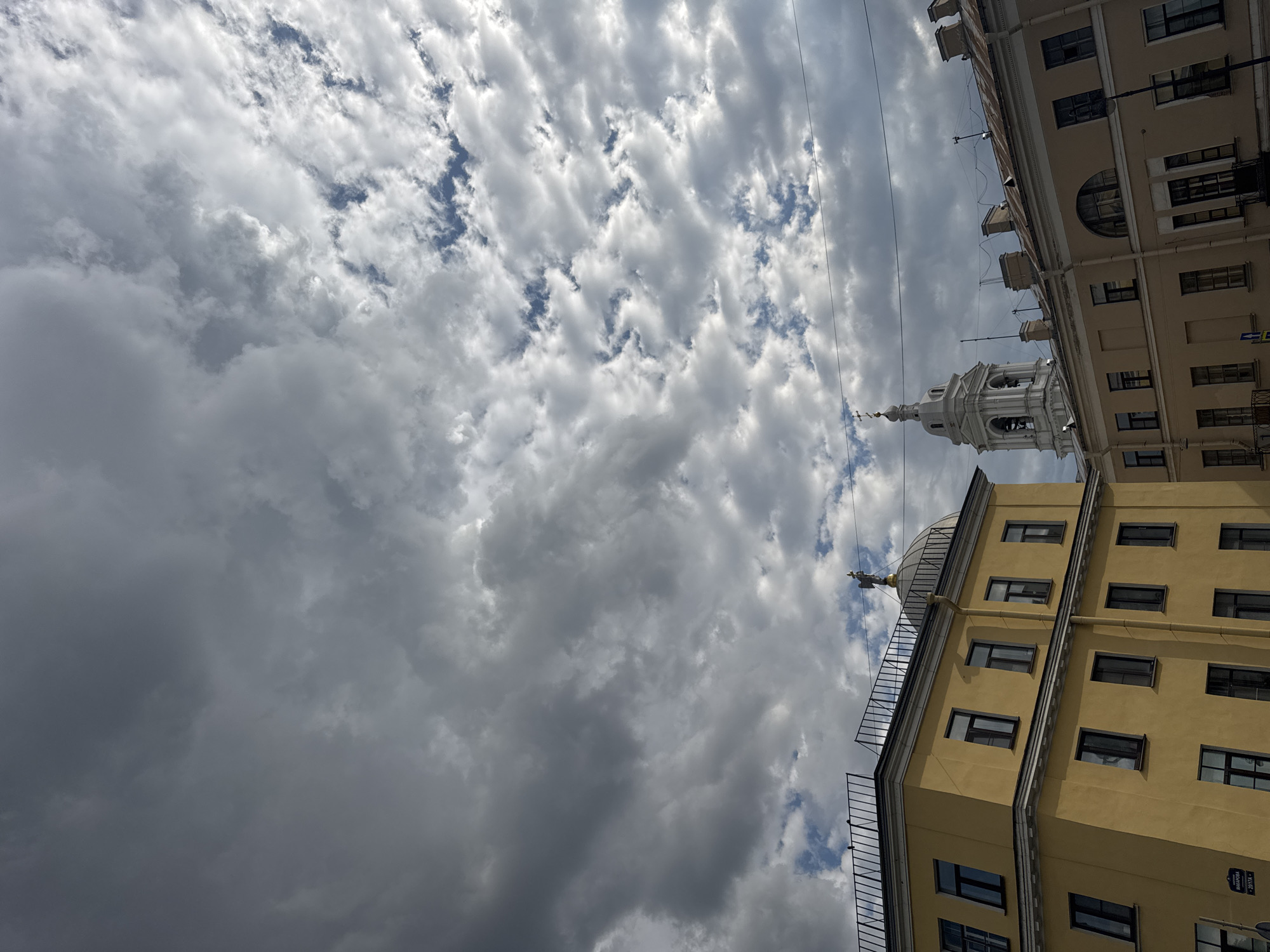 The image captures an upward-angled view of a city streetscape dominated by two buildings, a prominent bell tower, and a vast sky filled with clouds. The photograph is taken during daytime, under a dynamic sky that features a mix of bright, fluffy white clouds interspersed with patches of blue, and darker, heavier grey clouds, suggesting variable weather conditions.
In the foreground and to the right, a multi-story building with a warm yellow or ochre facade is visible. It features numerous rectangular windows with dark frames. The building has a flat roof adorned with a black metal railing, and at one corner, a small, light-colored dome topped with a golden, winged figure or finial can be seen. A dark blue street sign is partially visible on this building, bearing Cyrillic text that appears to read "МАЯКОВСКОГО" (Mayakovskogo), followed by numbers like "291074" or similar, indicating a street name in Saint Petersburg.
To the left and slightly behind the yellow building, a lighter, beige or cream-colored building rises, also featuring multiple windows, some rectangular and others with rounded arches on upper floors. Its architecture suggests an older, classic urban style.
Between these two structures, a distinct, white bell tower emerges, featuring several tiered sections with arched openings, culminating in a slender spire topped with an onion-shaped dome and a cross. This architectural detail is characteristic of Orthodox churches often found in Saint Petersburg.
Several thin, dark wires or cables stretch across the sky, traversing the scene horizontally and diagonally, adding a modern urban element to the traditional architecture below. The overall impression is one of a vibrant city, hinting at historical and architectural richness against a dramatic sky.