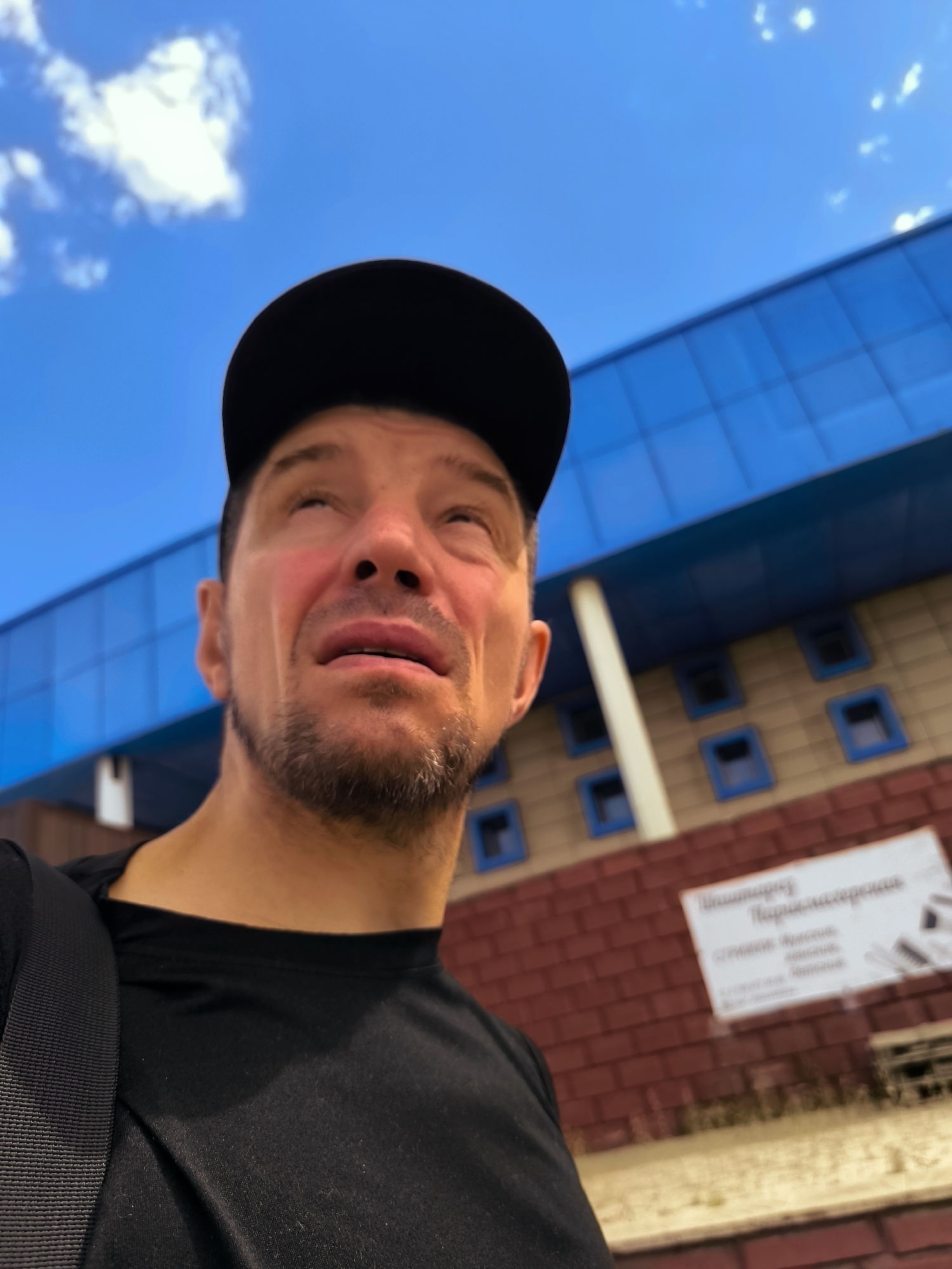 A man wearing a black baseball cap and a black t-shirt is looking upwards with his mouth slightly open and his eyes shut. He has a short beard and stubble. In the background, there is a building with blue glass windows on the upper section and brickwork on the lower section. A white sign with Russian text is affixed to the brickwork. The sky above is blue with some white clouds. A portion of a black bag strap is visible on the man's left shoulder. The image appears to be taken in an outdoor urban environment.