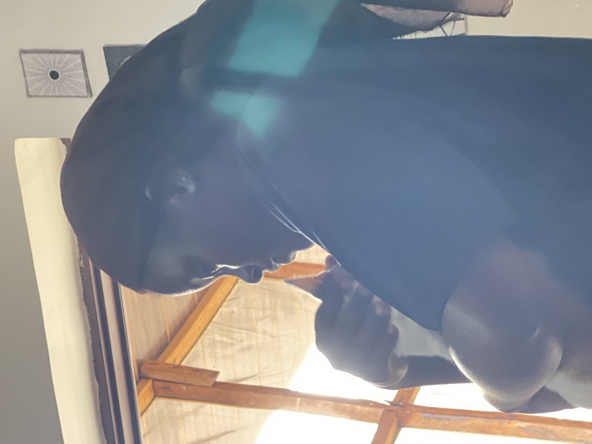 This media file features a close-up, Dutch-angled shot of a person wearing a black head covering and garment. The person's profile is visible, with their head tilted downwards as if looking at something out of frame. The lighting suggests it is daytime, with strong sunlight streaming in from what appears to be a window or opening, creating bright highlights and casting shadows. The background includes wooden beams, possibly part of a roof structure, and a light-colored, textured material that could be a tarp or fabric. On the wall in the upper left corner, there is a framed piece of art with a radial pattern. The overall impression is an indoor setting, possibly under construction or renovation, with natural light playing a significant role. There are no visible text elements in the image.