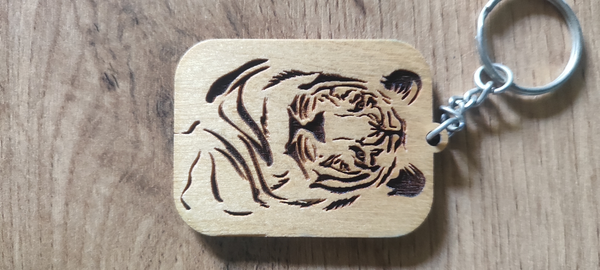 A light-colored wooden keychain with a carved tiger face is shown on a wooden surface. The tiger's face is detailed with carved stripes and features. A silver keychain ring and chain are attached to the top right corner of the keychain. The background is a wooden surface with visible grain. The scene is indoors, likely on a table or desk. There is no indication of location in Tashkent, Uzbekistan.