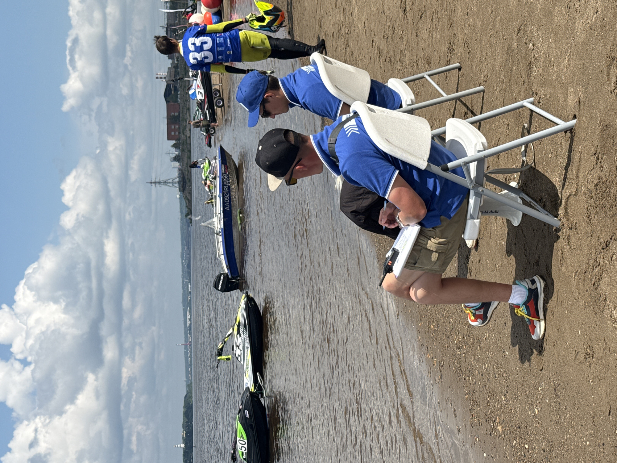 The image depicts a scene on the banks of the Kama River in the city of Perm, Russia, during a jet ski competition. In the foreground, two men are seated on white folding chairs on a sandy beach, facing away from the camera and towards the river. Both are wearing blue polo shirts, and the man on the left is wearing a black baseball cap and sunglasses, while the man on the right wears a blue baseball cap and sunglasses. Both have clipboards in their laps.
Several jet skis and a motorboat are visible on the water. The motorboat in the middle has the inscription "AA0921RUS59" on its side. Further back, another person in a blue and yellow racing suit with the number "33" on their back is walking away from the camera, carrying a jet ski helmet. The background shows a cityscape with industrial structures and a power line pylon, consistent with the industrial areas found along the Kama River in Perm. The sky is partly cloudy.