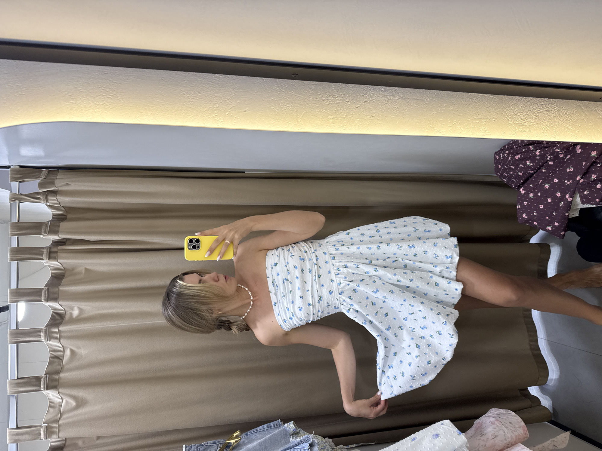 A woman, with blonde hair pulled into a braid, stands in a dressing room, taking a selfie with a yellow smartphone. She is wearing a white strapless dress with a blue floral print and a pearl necklace. The woman is holding up the hem of her dress with her left hand. Behind her, a beige curtain is drawn, and to her right, a tall, illuminated mirror reflects her image. Various clothing items, including a denim skirt and a purple floral-print fabric, are visible in the background. The dressing room appears to be located in a retail store.