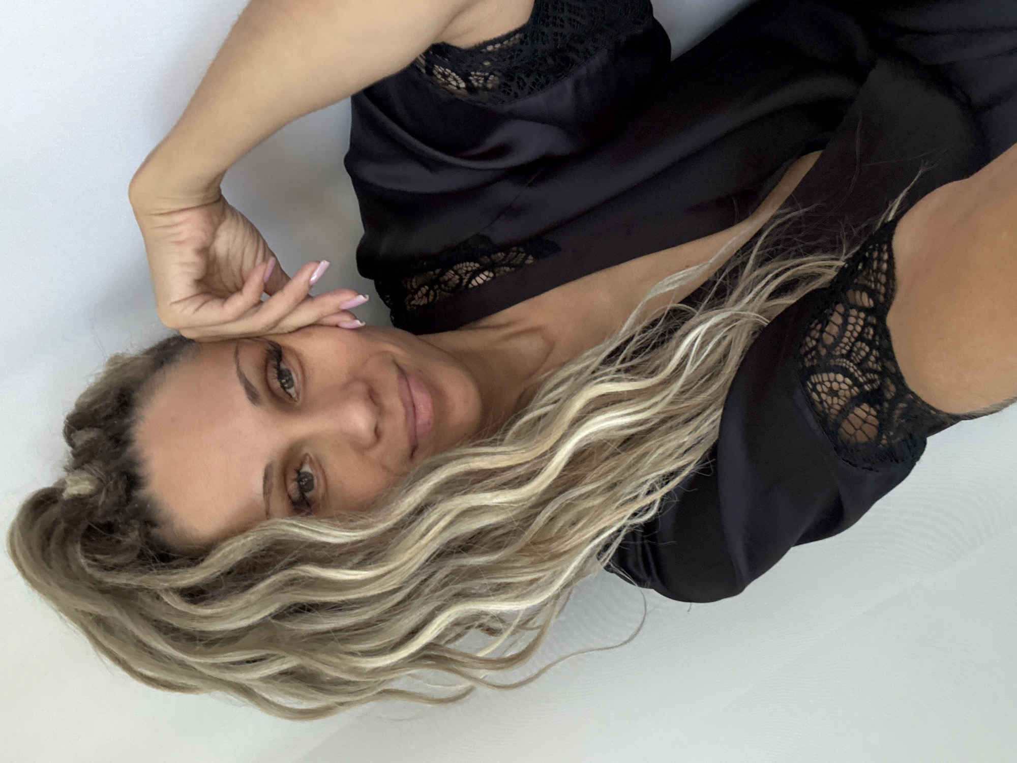 A woman with blonde dreadlocks pulled into a high ponytail is resting her hand on her forehead. She is wearing a black satin robe with lace trim on the sleeves and neckline. The background is a plain white wall. The image is a selfie. There is no information about the city of Rusanovo, Russia in this image.