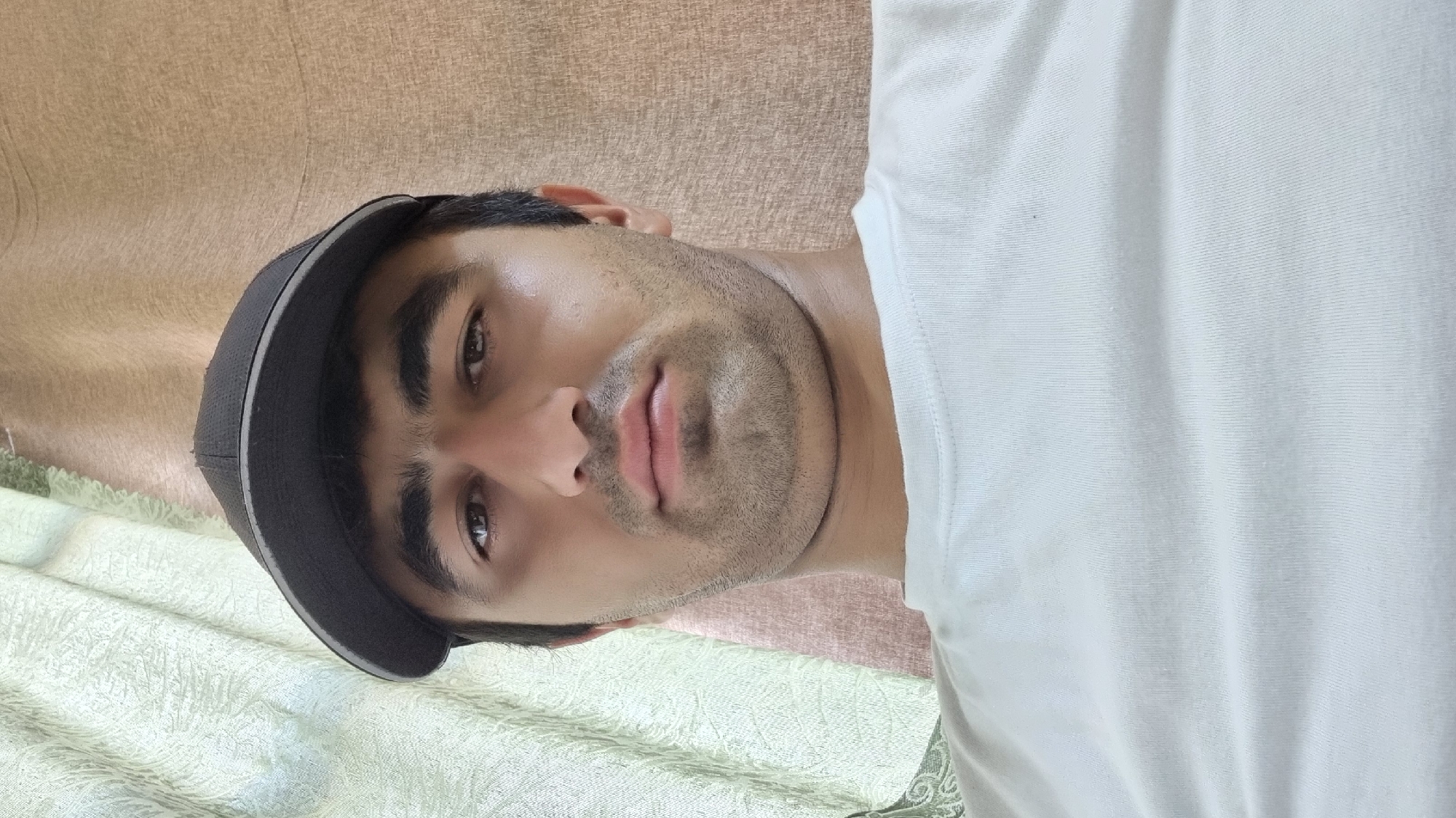 A close-up, head-on shot features a man wearing a dark baseball cap and a white t-shirt. He has dark hair, prominent eyebrows, and a light beard. The background consists of textured curtains in shades of light green and beige. The image appears to have been taken indoors. No specific location within the city of Ismoili Somoni, Tajikistan can be determined from the visual information provided.