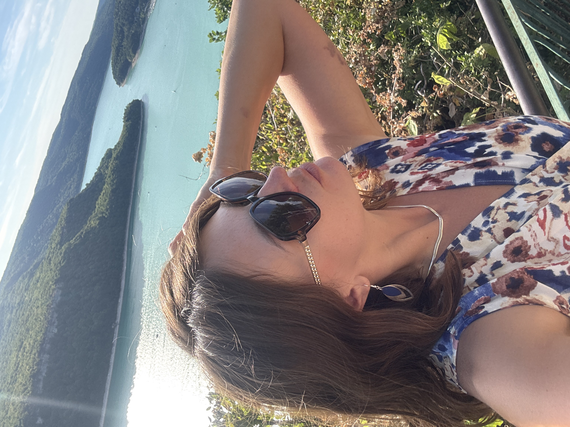 A woman is taking a selfie outdoors with a scenic body of water and forested hills in the background. She is wearing sunglasses, a silver necklace, and a floral print dress with blue, white, red, and brown patterns. Her hair is brown and she has a blue earring in her visible ear. She has her right arm behind her head, and her left arm is resting on her body. The background shows a large, turquoise lake with several boats, surrounded by lush green hills. The sun is shining brightly, creating a glare on the water and casting shadows. The image appears to be taken from a vantage point overlooking the lake. The location is identified as Moirans-en-Montagne, France.