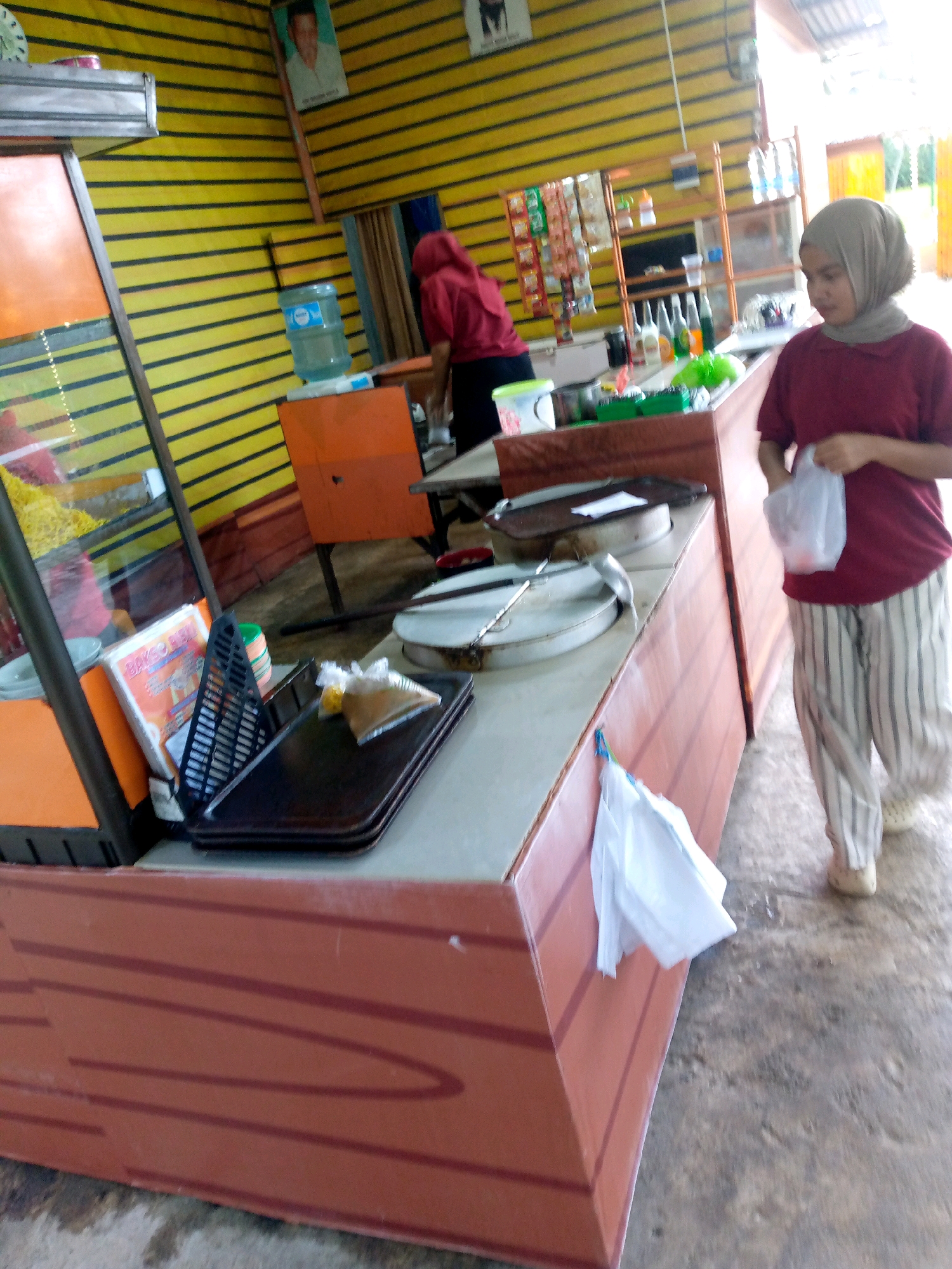 A young woman wearing a hijab and a maroon shirt with striped pants stands in front of a food stall in Gampongpaya, Indonesia. She is holding a plastic bag. The stall has yellow striped walls and a counter with various food items and cooking equipment. A man in a maroon shirt is visible behind the counter, and another woman in a red hijab is also present. The floor is concrete, and a wooden table is visible in the background. The scene depicts a typical street food vendor environment in Indonesia.