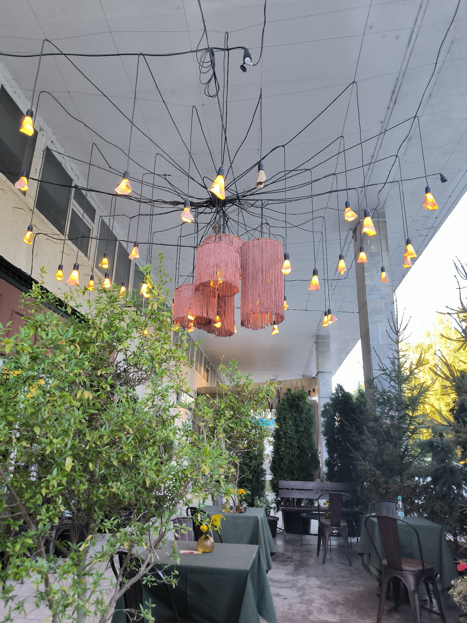 The image depicts an outdoor seating area with tables and chairs, suggesting a cafe or restaurant setting. The area is covered by a light-colored awning or roof, from which numerous decorative lights are suspended. These lights consist of exposed bulbs with orange-colored shades, arranged in a complex, web-like pattern of wires. In the foreground, lush green trees and bushes are visible, framing the seating area. Some tables are draped with dark green tablecloths, and one table has a small yellow floral arrangement in a glass vase. In the background, there are more trees and a bench. The overall atmosphere is one of a relaxed, possibly evening, outdoor dining or gathering space. The scene appears to be in Chișinău, Moldova, indicated by the presence of characteristic architecture and vegetation.