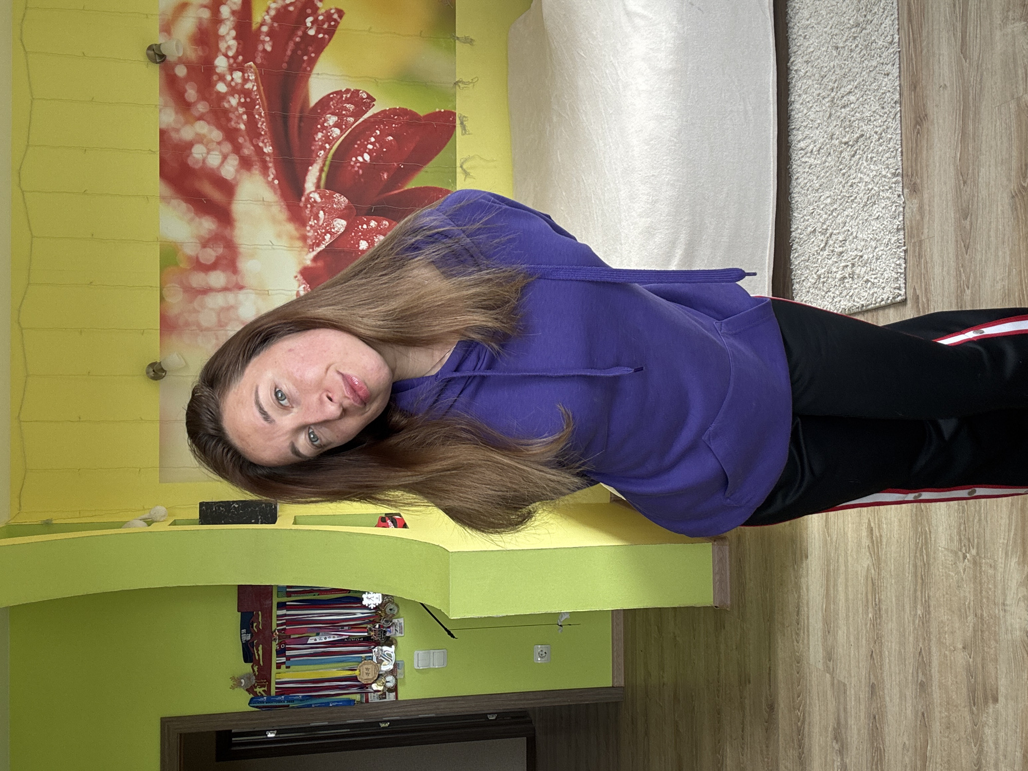 A woman is standing indoors in Прикамский, Russia. She has shoulder-length brown hair, fair skin, and blue eyes. She wears a purple hooded sweatshirt and black pants with a red and white stripe down the side. The background includes a couch, a rug, and a wall with a flower design. Below a ledge are medals and a TV. The walls are yellow and green. The floor is wooden.