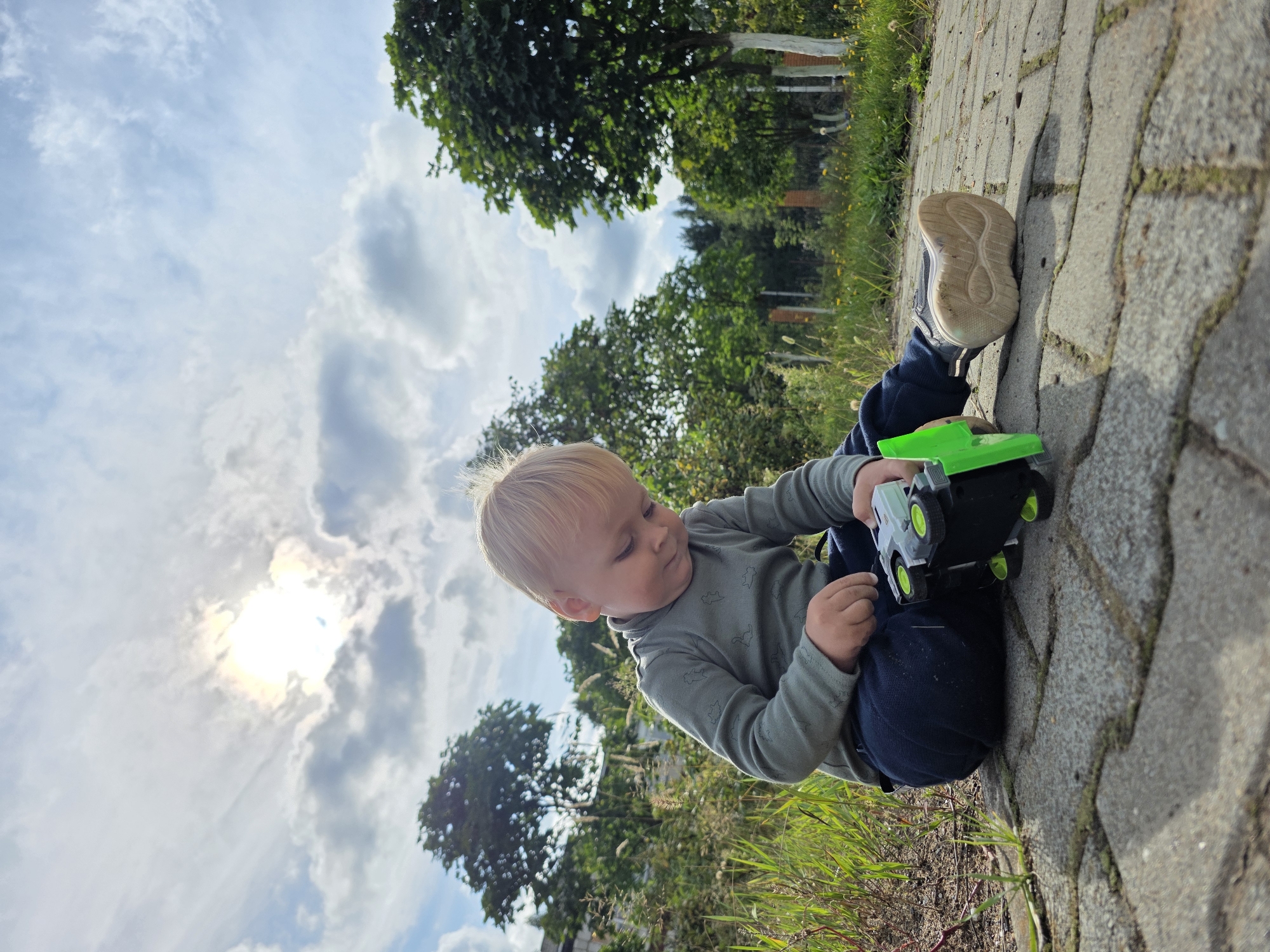 A young Caucasian boy with blonde hair sits on a cobblestone path outdoors, playing with a toy car. He is wearing a grey long-sleeved shirt with a dinosaur pattern and dark blue pants. The toy car is black and green. The background features green trees and grass under a partly cloudy sky with the sun shining brightly. The scene appears to be a park or residential area in Chaikovsky, Russia, given the presence of mature trees and the style of landscaping.