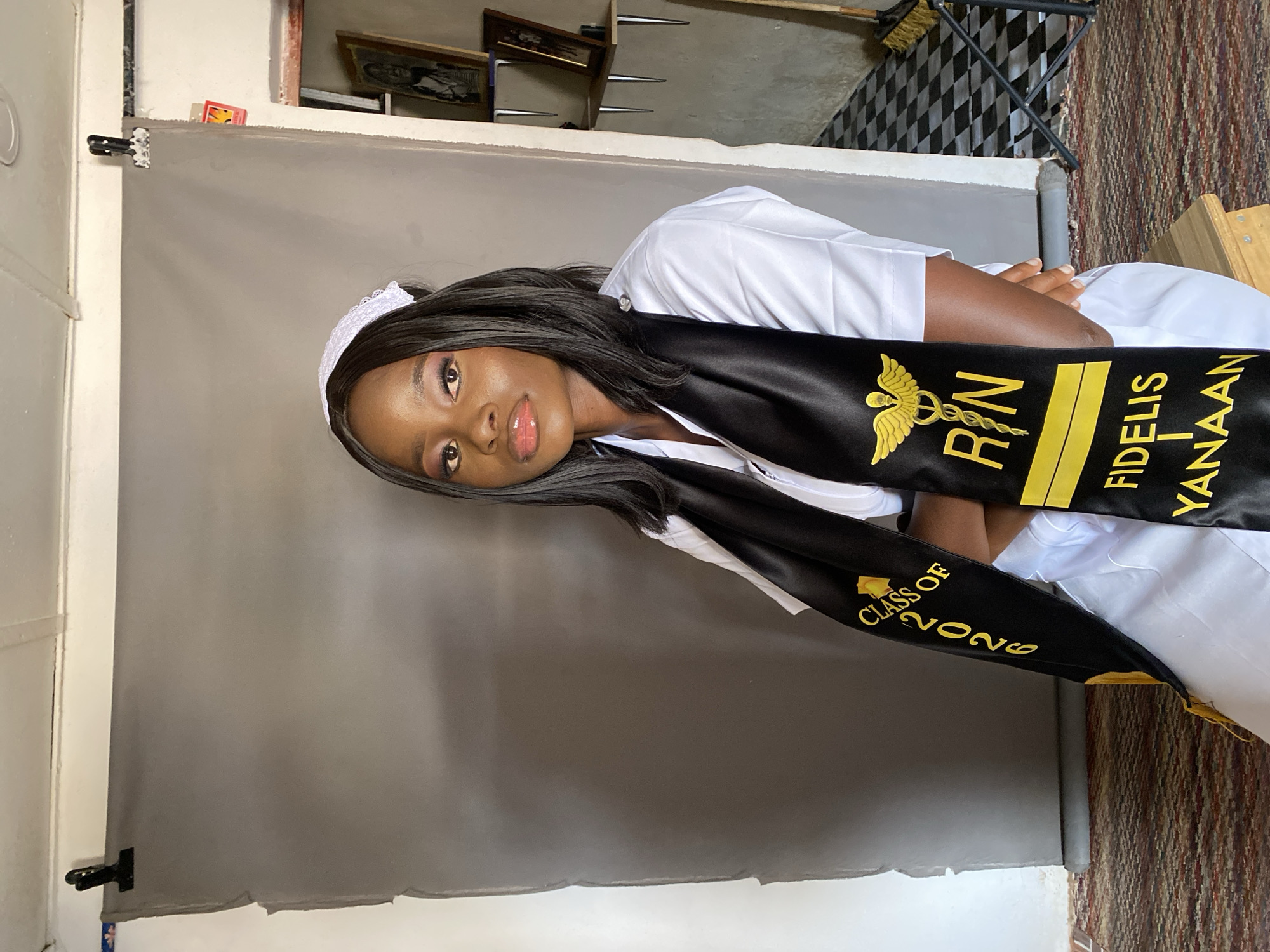 A young woman is captured in a portrait photograph, likely celebrating a graduation or professional milestone. She is positioned against a plain grey backdrop, which is visible on the left and bottom portions of the frame and held by clamps. The woman is wearing a white nurse's uniform, including a crisp white top and a white nurse's cap with a delicate lace trim. She also sports a black graduation stole draped over her shoulders.
The stole features prominent gold text and symbols. On her right side (viewer's left), a gold Caduceus symbol is displayed above the letters "RN" (Registered Nurse), followed by the name "FIDELIS YANAAN." On her left side (viewer's right), the text "CLASS OF 2026" is visible.
The woman has dark skin, long, straight dark hair, and is wearing subtle makeup, including pink lipstick and eye makeup. She looks directly at the camera with a calm and composed expression, her head slightly tilted, and her arms crossed.
In the background, behind and above the grey backdrop, elements of an indoor setting are visible, suggesting a makeshift studio. These elements include framed pictures, possibly shelves, a broom, and two distinct floor mats: one with a black and white checkered pattern and another with a woven, multi-toned texture. The overall scene conveys a sense of accomplishment and pride. The context provided indicates the photo was taken in Vom, Nigeria.