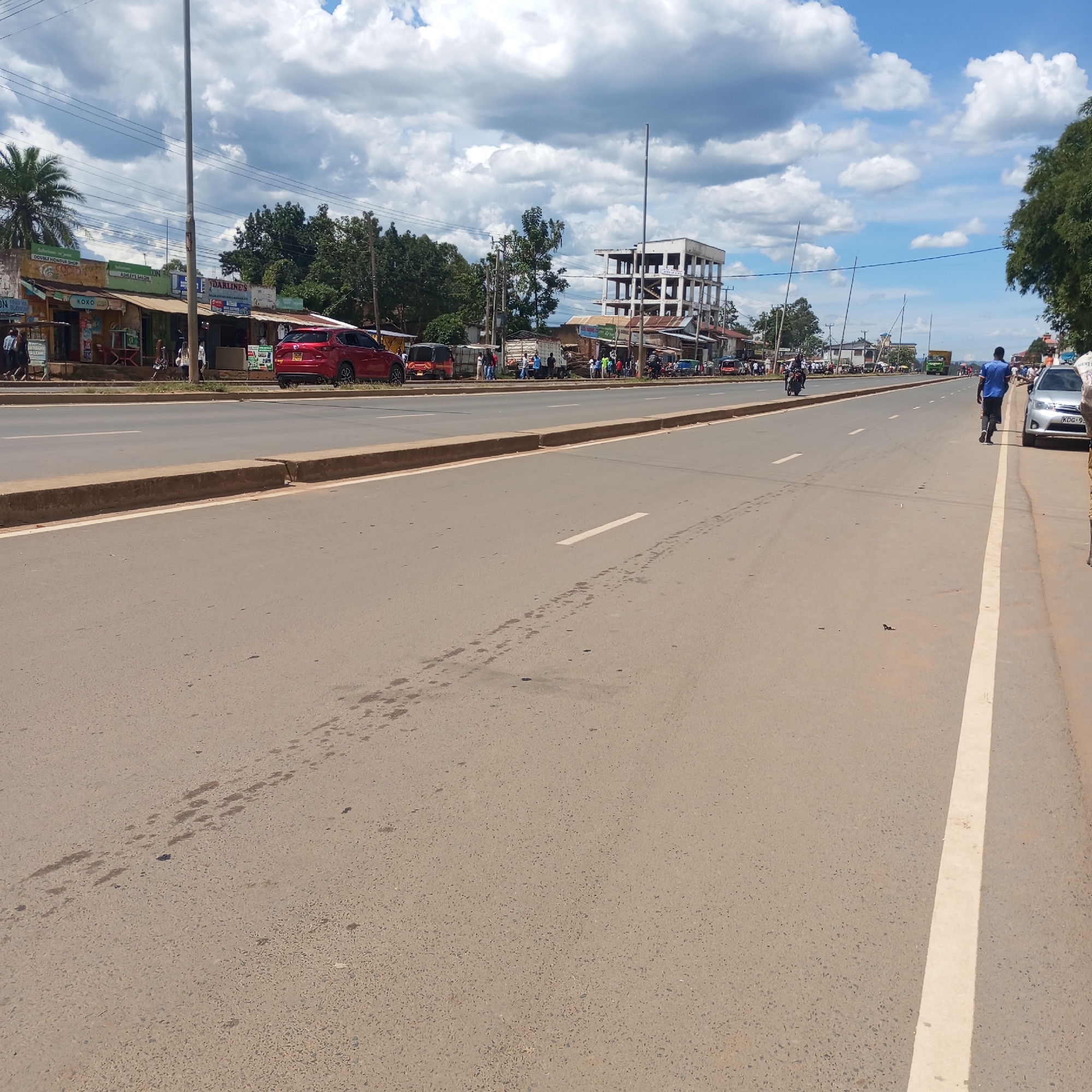 The image depicts a street scene in Sheywe ward, Kenya, under a sky with scattered clouds. The focus is on a wide, paved road with a central median and white lane markings. The asphalt shows signs of wear, with tire tracks and patches.
On the right side of the road, a man in a blue shirt and dark pants walks alongside the road. A silver car with license plate "KDG-9" is parked behind him.
On the left side, a row of buildings is visible, including small shops and commercial establishments with various signs and advertisements. A red car and a "tuk-tuk" are parked near the shops. People are standing or walking along the sidewalks in front of the buildings. In the background, a partially constructed building with a concrete frame stands out against the sky. Utility poles and wires run along the street.