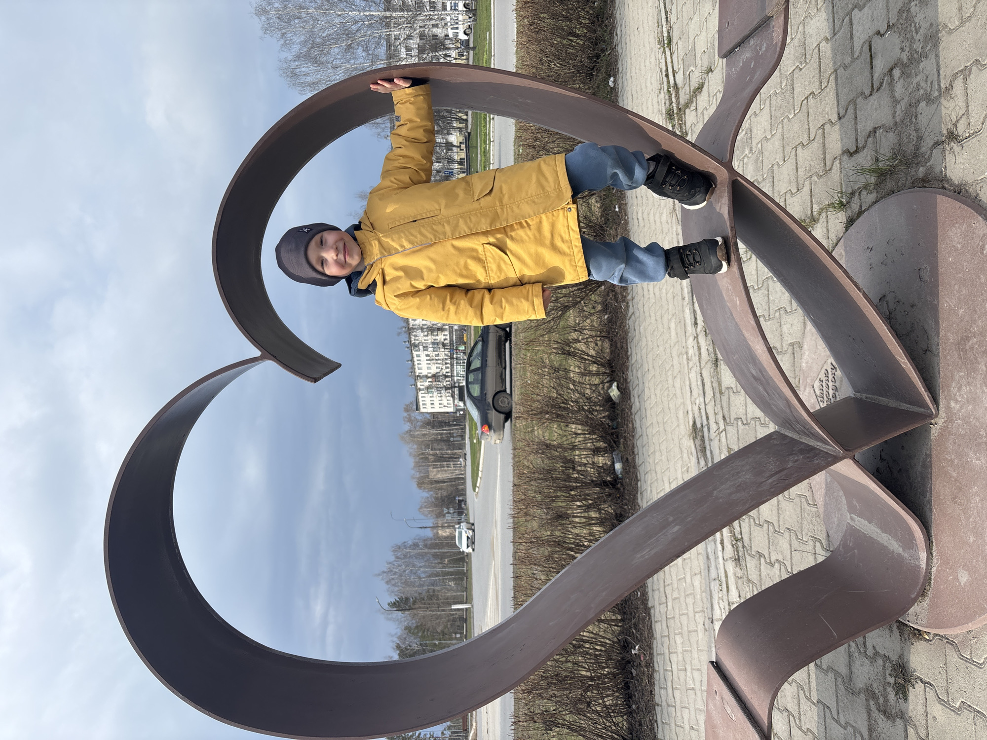 A young boy is playfully posing on a large, dark brown metal sculpture shaped like a heart, situated on a paved surface. The boy, dressed in a bright yellow winter coat, a grey beanie with a white emblem, blue sweatpants, and black sneakers, stands with one foot on each side of the heart's upper curve. He holds onto the top edge of the sculpture with both hands and looks towards the camera with a happy, smiling expression.
The scene is an outdoor urban or suburban environment, likely a public space or park adjacent to a road. In the background, a street is visible with a dark grey car and a white car, bordered by numerous bare trees, suggesting late winter or early spring. Distant multi-story buildings can be seen beyond the trees and road. The sky is partly cloudy with visible patches of blue.
It is daytime, with clear natural lighting. The boy's warm clothing indicates cool or chilly weather. Faint, weathered script is visible on the base of the sculpture, which appears to include text related to "Chaikovsky" or "город Чайковский" (City of Chaikovsky), aligning with the location context of Chaikovsky, Russia. The image captures a moment of cheerful interaction between the child and the public art installation.