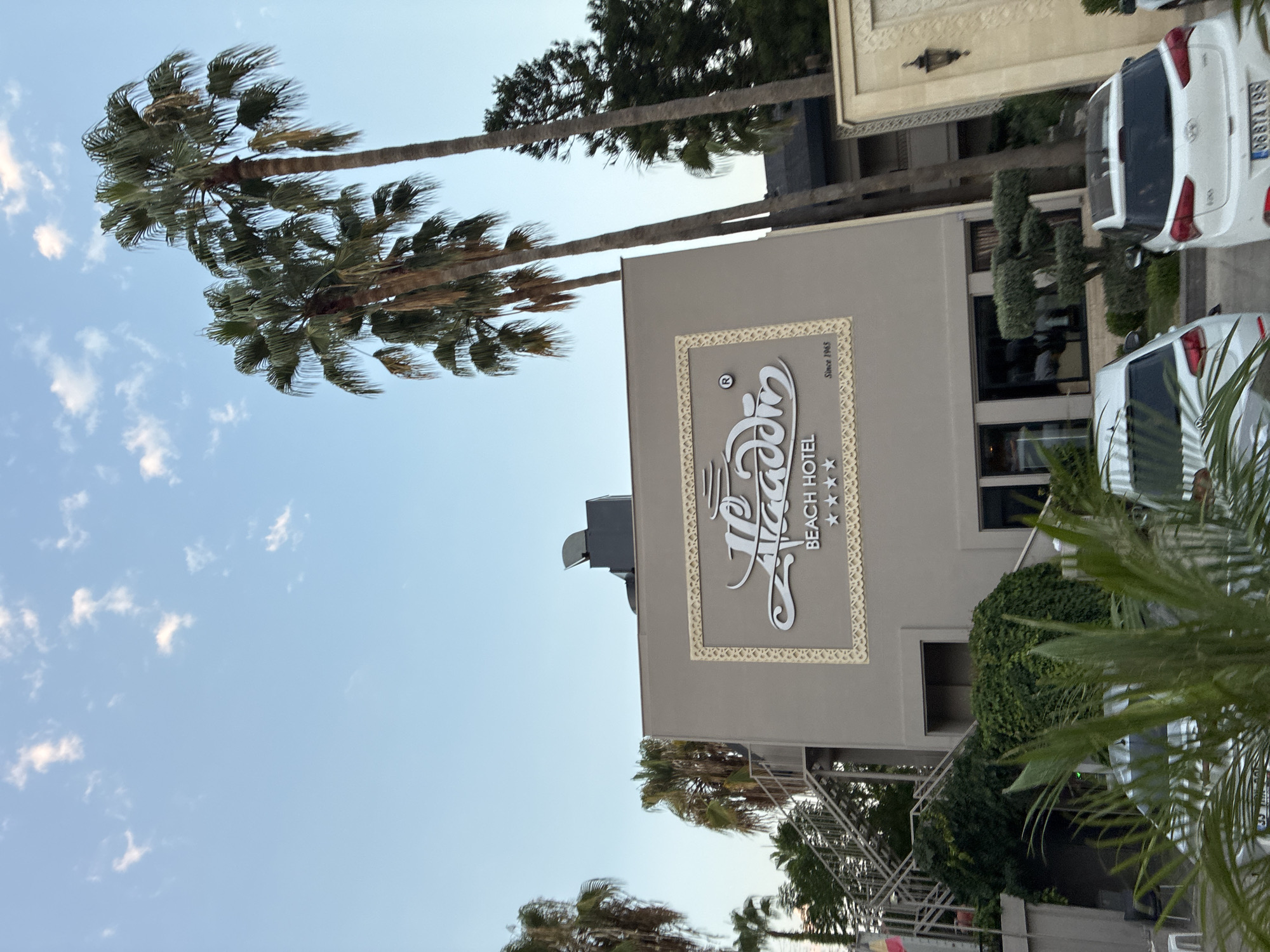 The image shows the front of the Aladdin Beach Hotel in Alanya, Turkey. The hotel has a tan facade with a large, ornate sign that reads "Aladdin Beach Hotel" in white lettering. The sign is surrounded by a decorative border and features three stars beneath the hotel name. To the left of the entrance, there is an outdoor staircase covered in greenery. Several palm trees are visible on the right side of the hotel. In the foreground, two white cars are parked. The sky is blue with scattered white clouds.