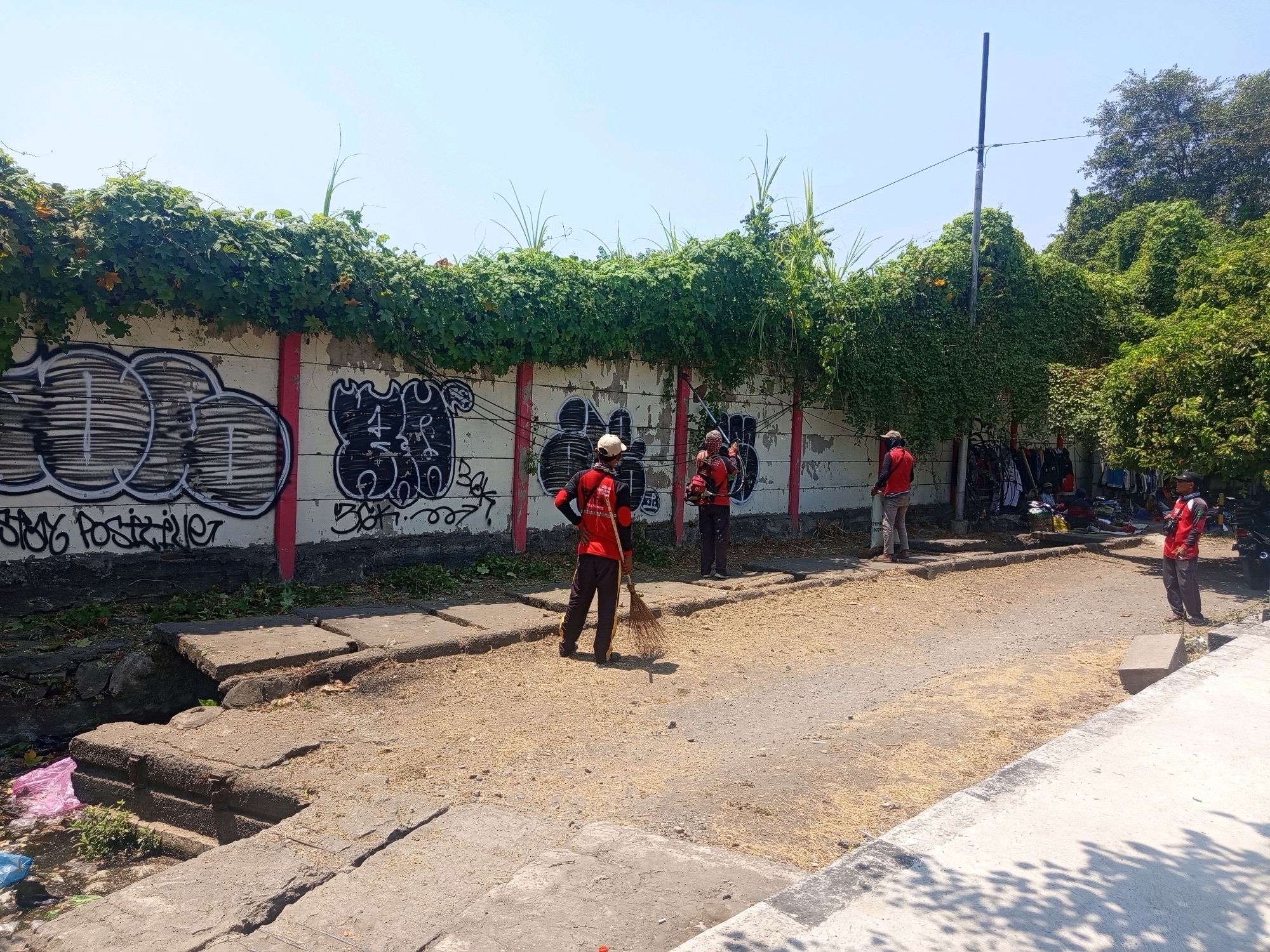 Four individuals in red uniforms are working along a concrete wall covered in green ivy and graffiti. Two individuals are sweeping the ground, one with a broom and another with a leaf blower. The area appears to be an outdoor public space with a dirt path and concrete walkways. Several motorcycles and what appear to be market stalls are visible in the background. The scene suggests a cleanup or maintenance effort is underway. The image may have been taken in Semarang, Indonesia, given the context provided.