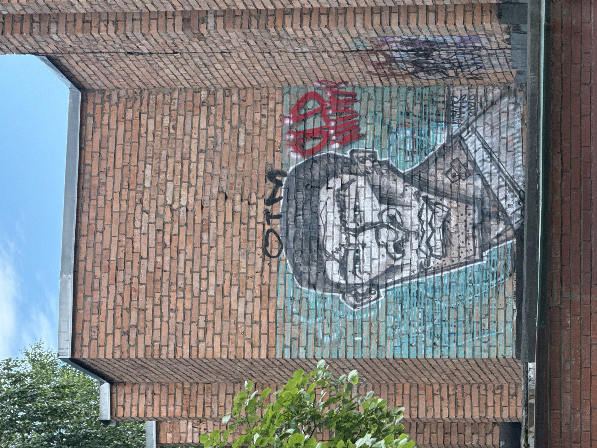 A graffiti mural of a man's face is spray-painted on the side of a brick building in Votkinsk, Russia. The mural is on a turquoise background and features the man's face in black and white. Above the face, the letters "OTM" are spray-painted in black. To the right of the mural, there are red spray-painted letters. The building is made of red brick and is partially covered by a tree with green leaves on the left side of the image. The sky is blue and visible above the building.