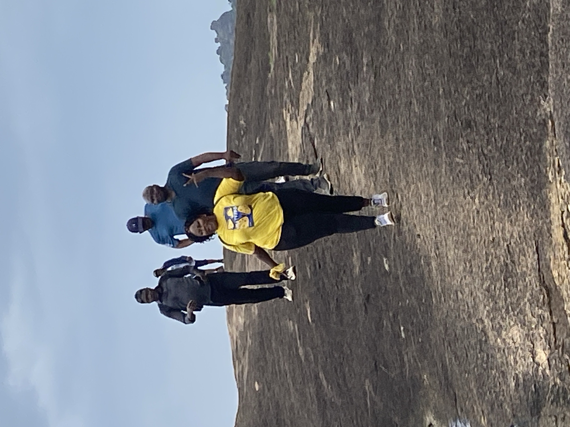 Four people are posing for a photo in front of a large rock formation. The people are dressed in casual clothes. The person in the center is wearing a yellow shirt and is standing with their arms outstretched. The sky is blue. The location is Ganawuri, Nigeria.