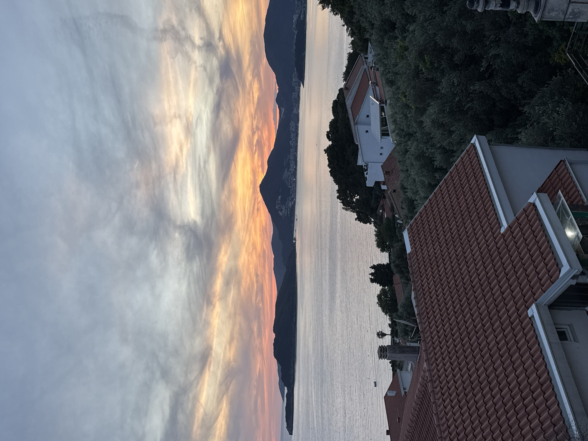 The image depicts a sunset view in Sveti Stefan, Montenegro, looking toward the mountains across the water. The sky exhibits a blend of gray and orange hues, indicating the sunset. The landscape is divided by water. The left side shows a mountain range and the right shows the edge of a building with red tile roofing and trees. The image orientation is rotated 90 degrees counterclockwise.