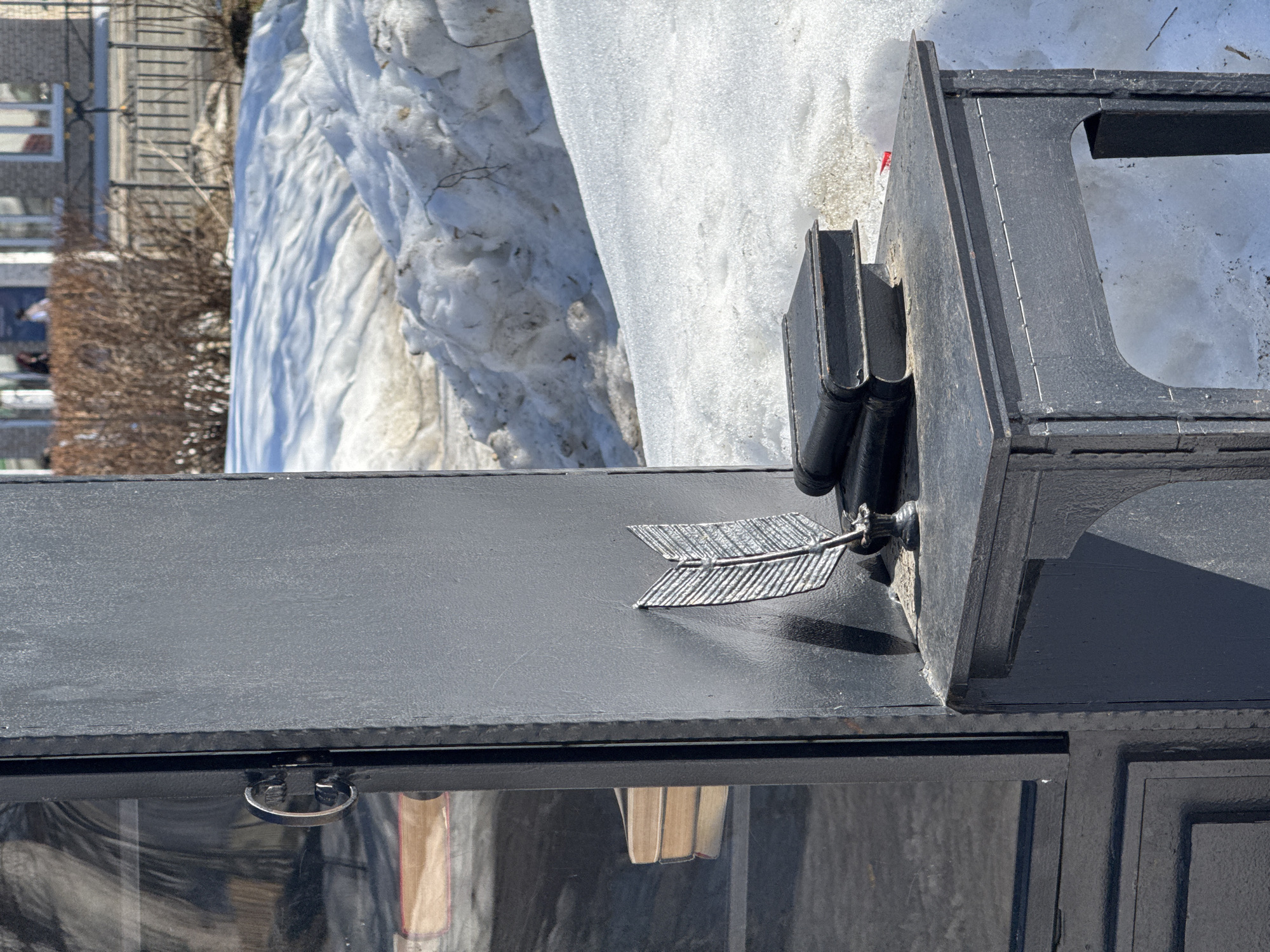 An outdoor, dark grey/black metal structure, likely an art installation or public mini-library, stands amidst melting snow in Chaikovsky, Russia. The structure features a glass-fronted cabinet on the left, secured by a silver-colored handle, revealing multiple light-spined books stacked horizontally inside. An attached, smaller dark grey/black metal table extends to the right, upon which sit a stack of two dark-bound books, a silver-colored metal quill pen, and a black metal inkwell. The ground is covered with patchy, dirty white and brown melting snow, with visible brown earth. In the blurred background, bare brown bushes, a dark metal fence, and light-colored buildings with white windows are visible. Several out-of-focus individuals can be seen near the buildings. The scene is illuminated by bright sunlight.