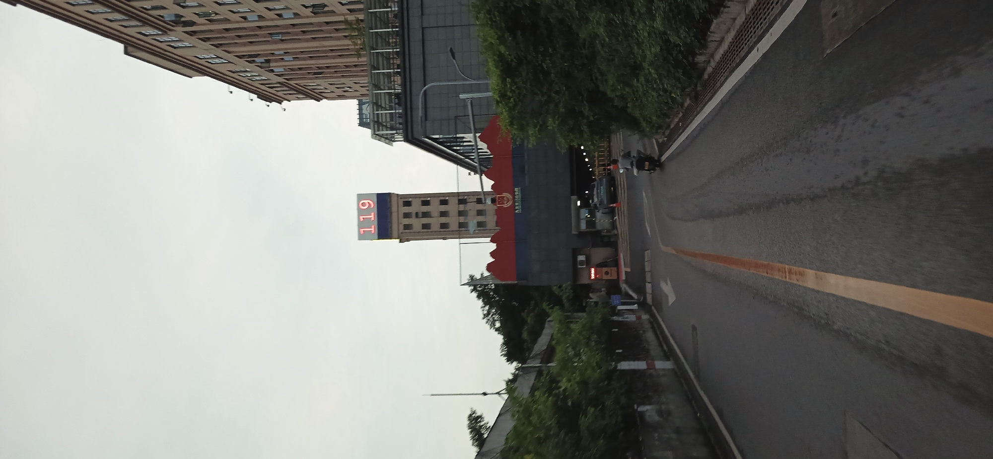 The image depicts a street scene in Jiulongpo District, China. In the foreground, a road with a yellow line running along the edge is visible. A building with the number "119" displayed vertically in red is seen in the background. A person on a motorbike is moving along the road. There are other buildings visible. The sky is overcast.