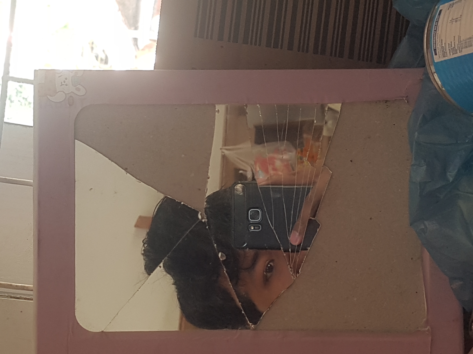 The image shows a person taking a selfie in a cracked mirror. The mirror is framed by a pink border and appears to be part of a vanity. The person's face is partially visible, along with their hand holding a black smartphone. Behind the mirror, there is a window with natural light streaming in, suggesting an indoor setting. The background also reveals some household items, including what appears to be a blue trash bag and a cylindrical container. The context provided about the location being Tinatar, Indonesia, does not offer any visual cues within the image itself.