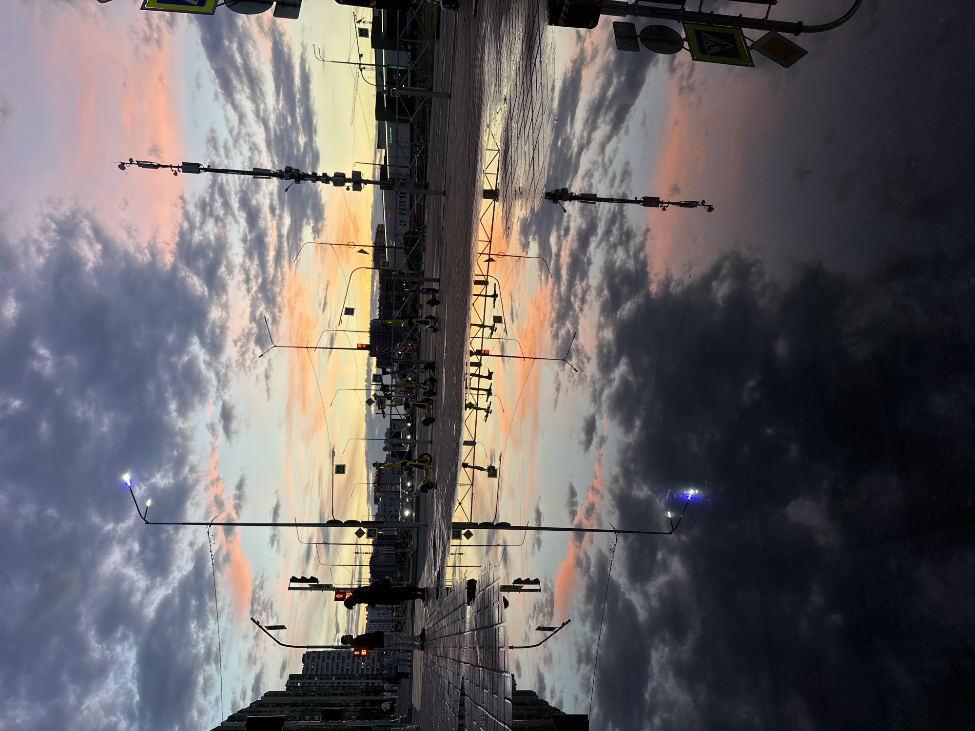 In the city of Unknown, Unknown, a street scene is captured at dusk, with a dramatic sunset reflected in a large puddle on the paved sidewalk. The sky is filled with a gradient of colors, from deep grays and blues in the clouds to vibrant pinks and oranges near the horizon.
Silhouettes of two individuals are visible standing on the wet pavement. One person, facing left, is wearing what appears to be casual clothing. The other, facing right, is also in silhouette. In the background, streetlights illuminate the scene, with one exhibiting a blue light. Across the street, parked electric scooters are lined up, and traffic lights are visible. Further in the distance, the outlines of buildings and construction cranes are discernible against the colorful sky. The reflection in the puddle mirrors the sky and the streetlights, creating a symmetrical and somewhat surreal effect. The overall impression is one of a tranquil urban evening, enhanced by the natural spectacle of the sunset.