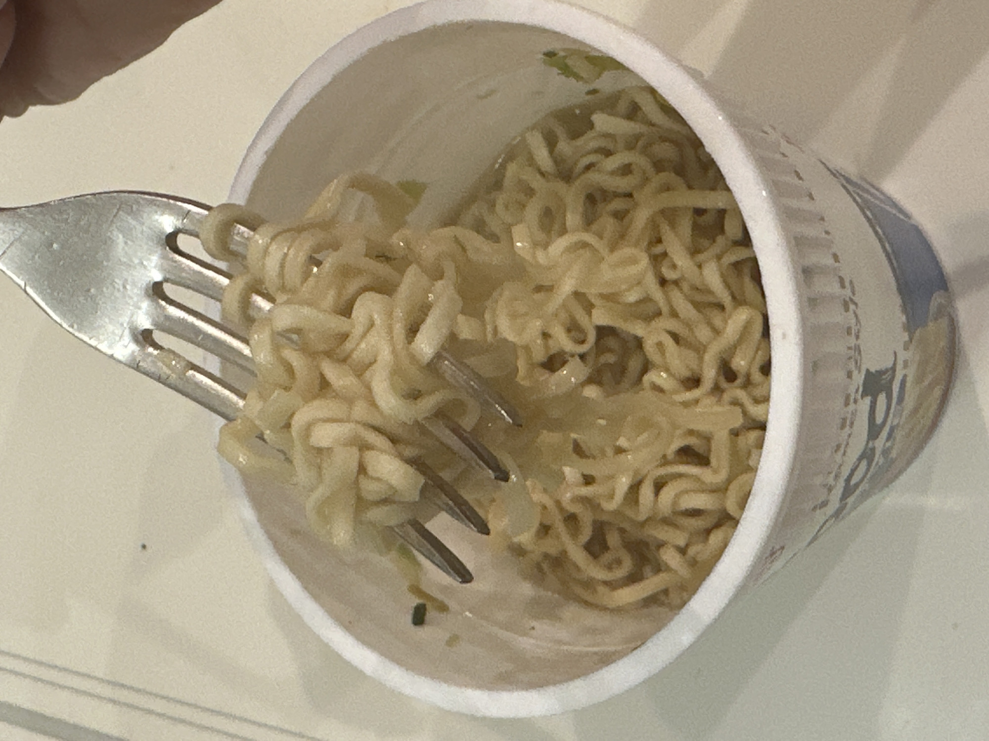 A close-up, eye-level image depicts a white, disposable cup containing rehydrated instant noodles and a light-colored broth. A silver metallic fork is positioned inside the cup, with its tines submerged and lifting a substantial portion of wavy noodles from the liquid. The upper right quadrant of the image shows a portion of a human hand, primarily the thumb and forefinger area, gripping the handle of the fork. Small green fragments, consistent with dehydrated vegetables, are visible within the broth and intermingled with the noodles. The exterior of the cup features faint blue and potentially other colored graphics near its base. The cup rests on a uniform, light-colored surface, possibly a countertop or table. The background consists of a blurred, lighter-toned surface. The specific location in the city of London, United Kingdom, cannot be determined from the visual evidence presented.