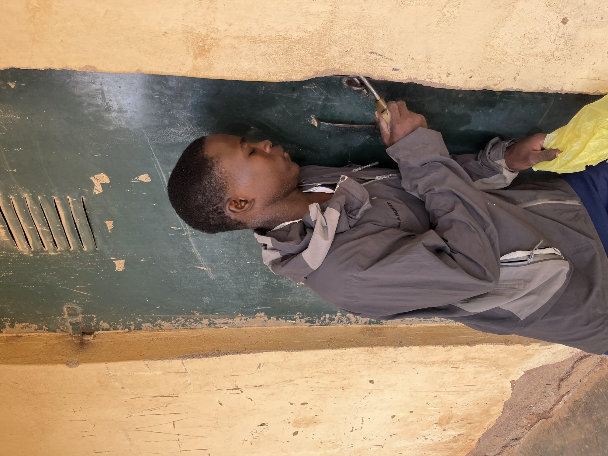 A young Black male wearing a grey hooded jacket and blue shorts is shown from the chest up, standing in a doorway with a dirty yellow wall to his left and a dark green door to his right. He is holding a small tool in his right hand and a yellow plastic bag in his left, and appears to be inspecting or attempting to open the door. The door has a metal hinge and a visible gap. The wall on the left has graffiti. The background shows a sliver of the interior, with horizontal vents. The scene suggests an outdoor, possibly urban or semi-urban, environment.