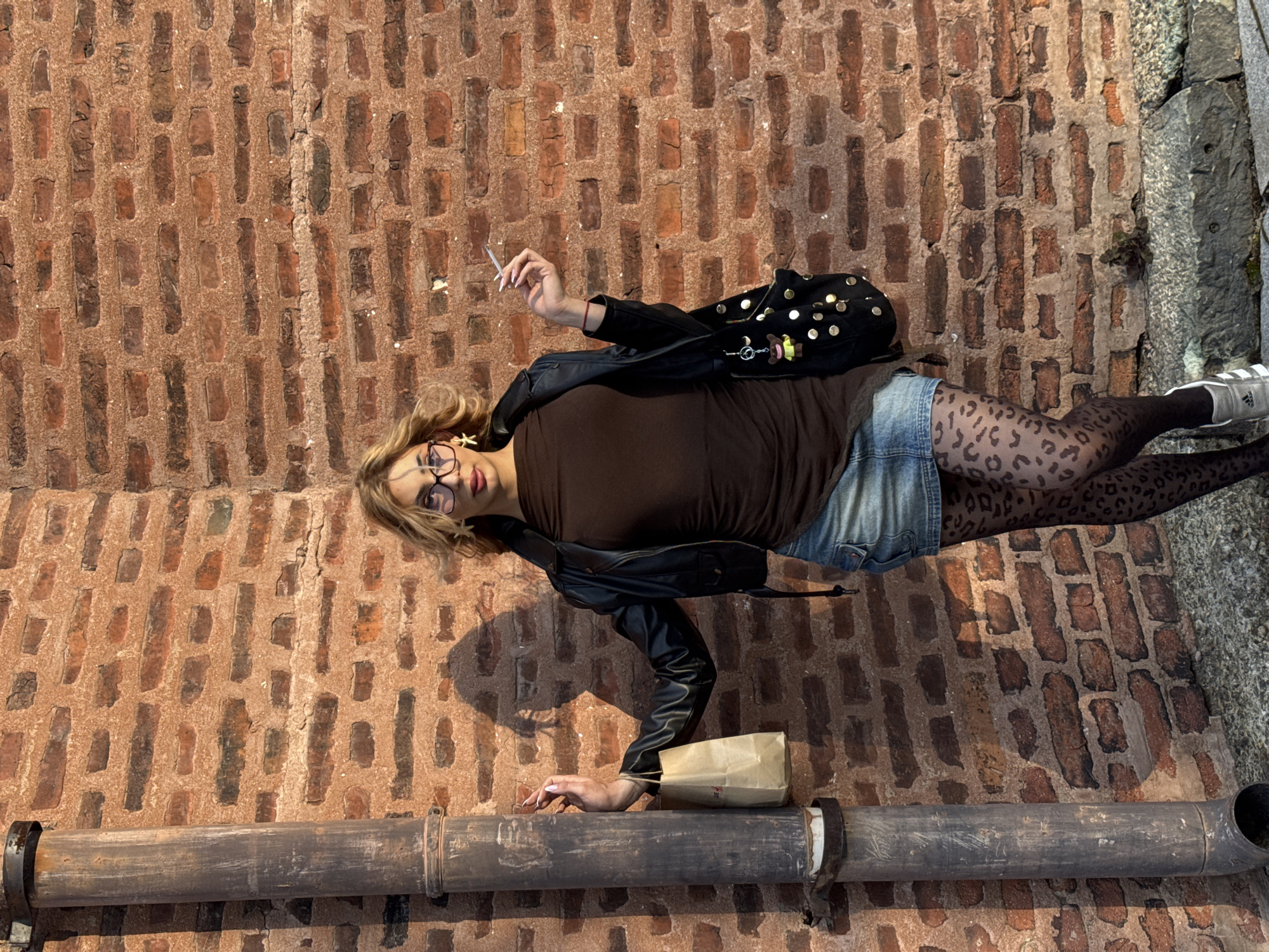 This image, taken in Sofia, Bulgaria, portrays a young woman lying horizontally against a reddish-brown brick surface, likely a wall. She has blonde hair, wears glasses, and looks directly towards the viewer. She is dressed in a brown top, a black leather jacket adorned with gold studs and decorative charms (including a small teddy bear), denim shorts, and black leopard-print stockings, finished with light-colored Adidas sneakers.
In her right hand, she holds a lit cigarette with white-painted nails. Her left hand is resting near a small, crumpled brown paper bag, which is partially supported by a large, weathered metal pipe running horizontally across the bottom of the frame. The background is dominated by the old, irregular brickwork, with a rough concrete or stone structure visible on the far right side of the image. The scene is brightly illuminated by natural daylight, casting some distinct shadows. Visible text includes the Adidas logo on her shoe. The paper bag has faint, partially obscured text that appears to start with "DO NOT", and the brick wall displays some faded, illegible markings.