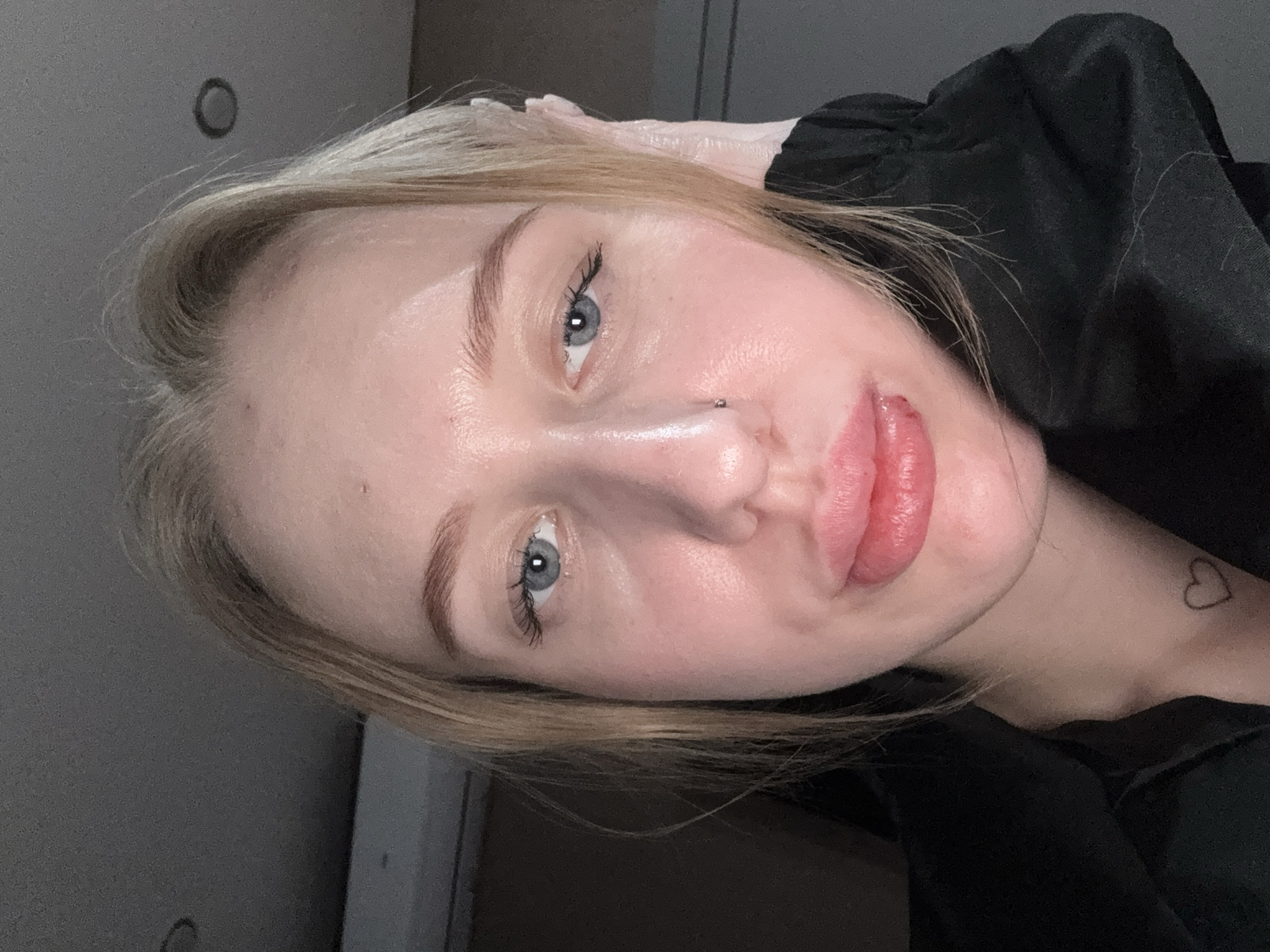 An indoor, close-up portrait photograph depicts a young adult female, presumed to be in Azov, Russia. The subject has light skin, blonde hair, and blue eyes. A small metallic stud piercing is present in her left nostril. Her lips appear full, with a visible reddish-brown mark or scab on the center of her lower lip. A small, outlined heart tattoo is located on her left inner forearm, visible below the neckline of her dark, possibly black, garment. Her left hand is partially visible on the right side of the frame, appearing to rest against her head or hair. The background is a dimly lit, indistinct interior, featuring a dark ceiling with a circular recessed light fixture visible in the upper right portion of the frame. The subject is looking directly towards the camera.