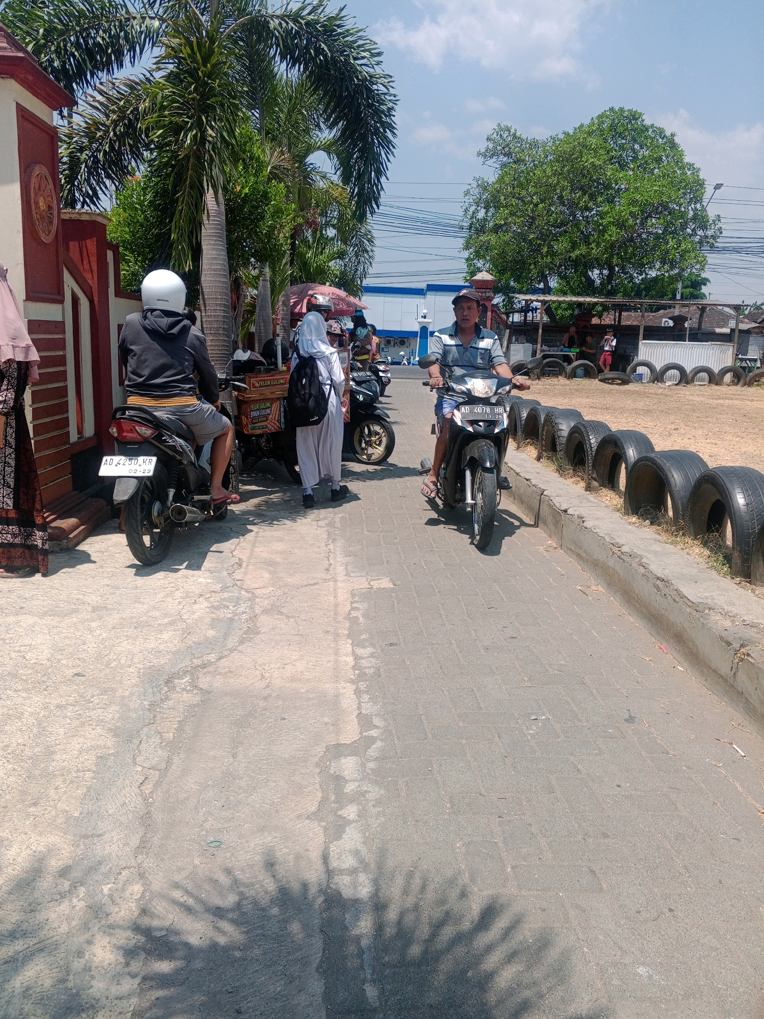 In the city of Giritirto, Indonesia, a man wearing a striped shirt and shorts rides a black motorcycle down a paved road. He is wearing a baseball cap. To his left, another motorcycle is parked, and a person wearing a white hijab and a backpack is standing near it. Another individual, wearing a dark hoodie and white helmet, is seated on a parked motorcycle. In the background, palm trees and other foliage are visible, along with a building and a row of stacked tires. The sky is clear and blue with some clouds.