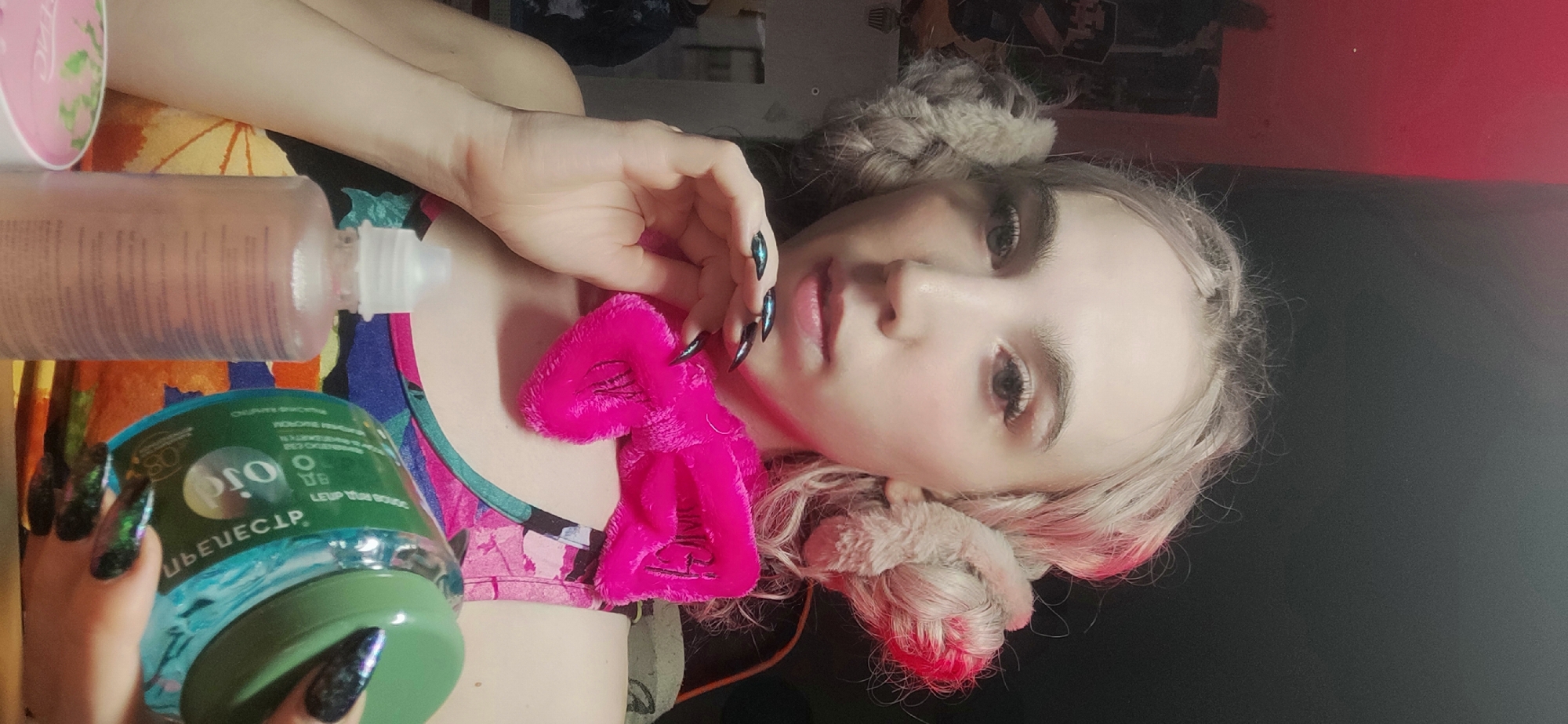 A woman lies on her back, wearing a pink bra and a pink headband with a bow. She has long, claw-like nails with an iridescent finish. She is holding a green container of "G ELSEVE CLIP" and a clear bottle with a white top. Her hair is braided and has earmuffs on. A pink container is also visible in the upper left corner. The location is Chaikovsky, Russia.