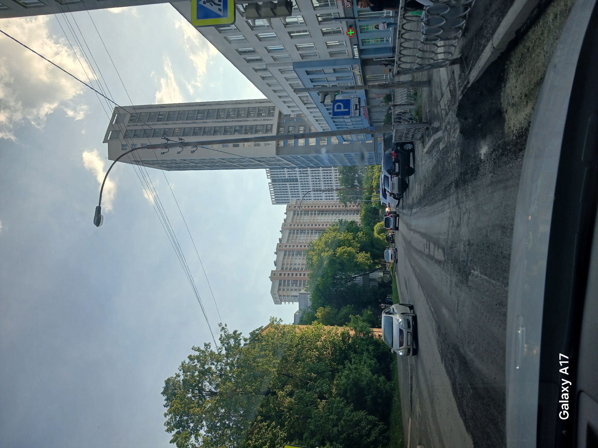 The image, taken from inside a vehicle, shows a street scene in Yekaterinburg, Russia. On the right side of the image, there are cars parked alongside the road, and other vehicles are moving along the street. On the left side, there are tall buildings, a sidewalk, trees, a streetlamp, and overhead power lines. A parking sign is visible near one of the buildings. The sky is partly cloudy.