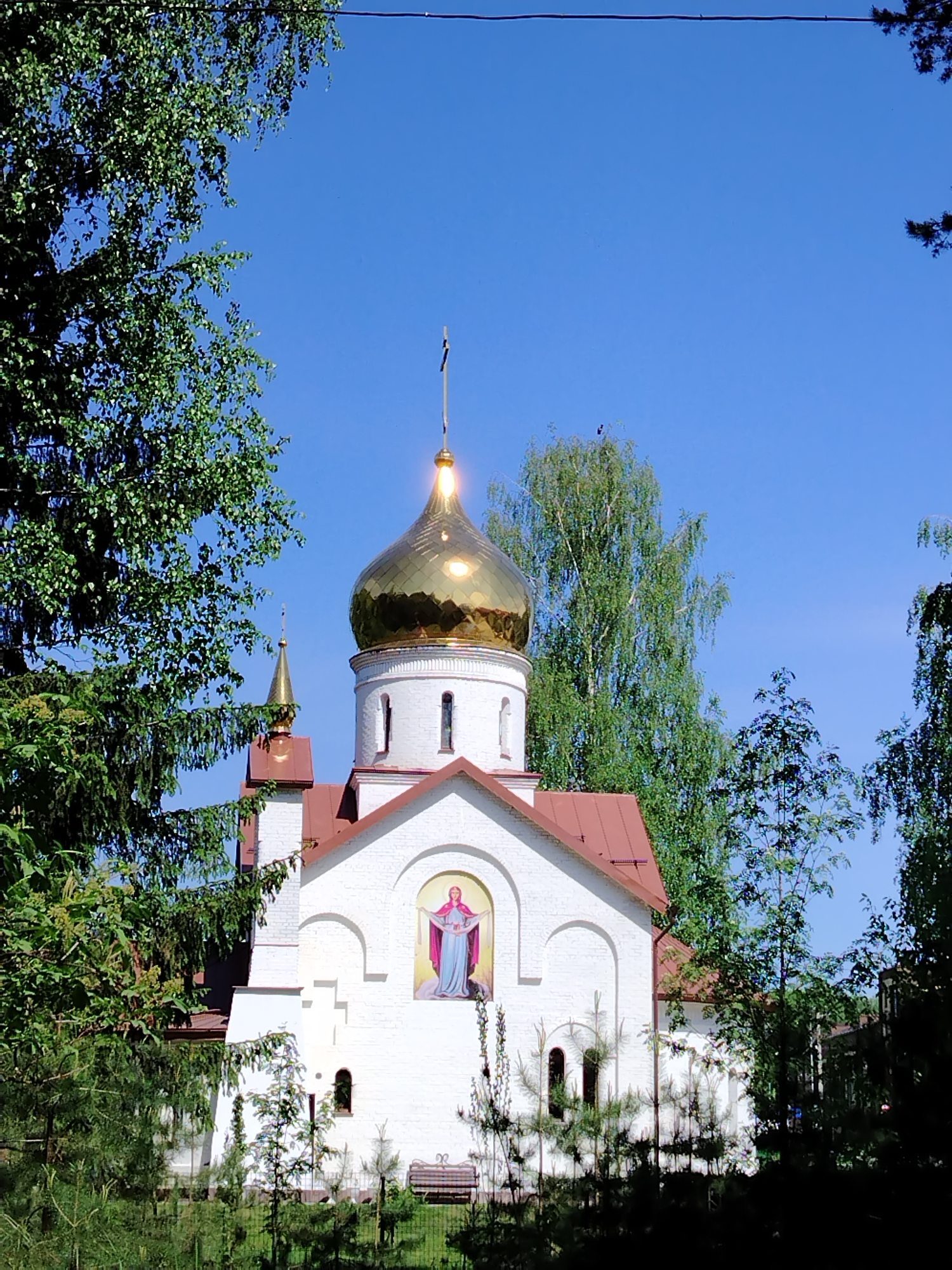 The image depicts a white brick church with a golden dome and a red roof, framed by trees against a clear blue sky. An icon of a figure in red and blue robes is positioned in an arched recess above the entrance. In front of the church, there's a fence, small trees, and a bench. The photograph was taken in Noviy, Russia.