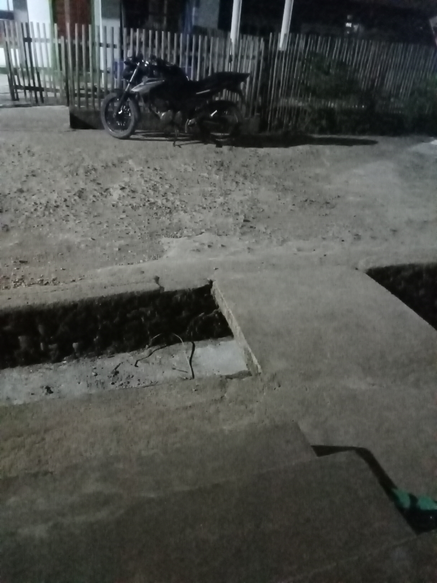 The image shows a dark scene outside, possibly at night or in low light conditions, featuring a motorcycle parked in front of a wooden fence. The ground is a mix of gravel and concrete. In the foreground, there's a concrete structure with a dark trench or opening. The overall environment appears to be an outdoor urban or semi-urban setting. Specific location details for Setia Jaya, Indonesia, are not discernible from the visual information provided in the image. There are no people or discernible actions or interactions depicted.