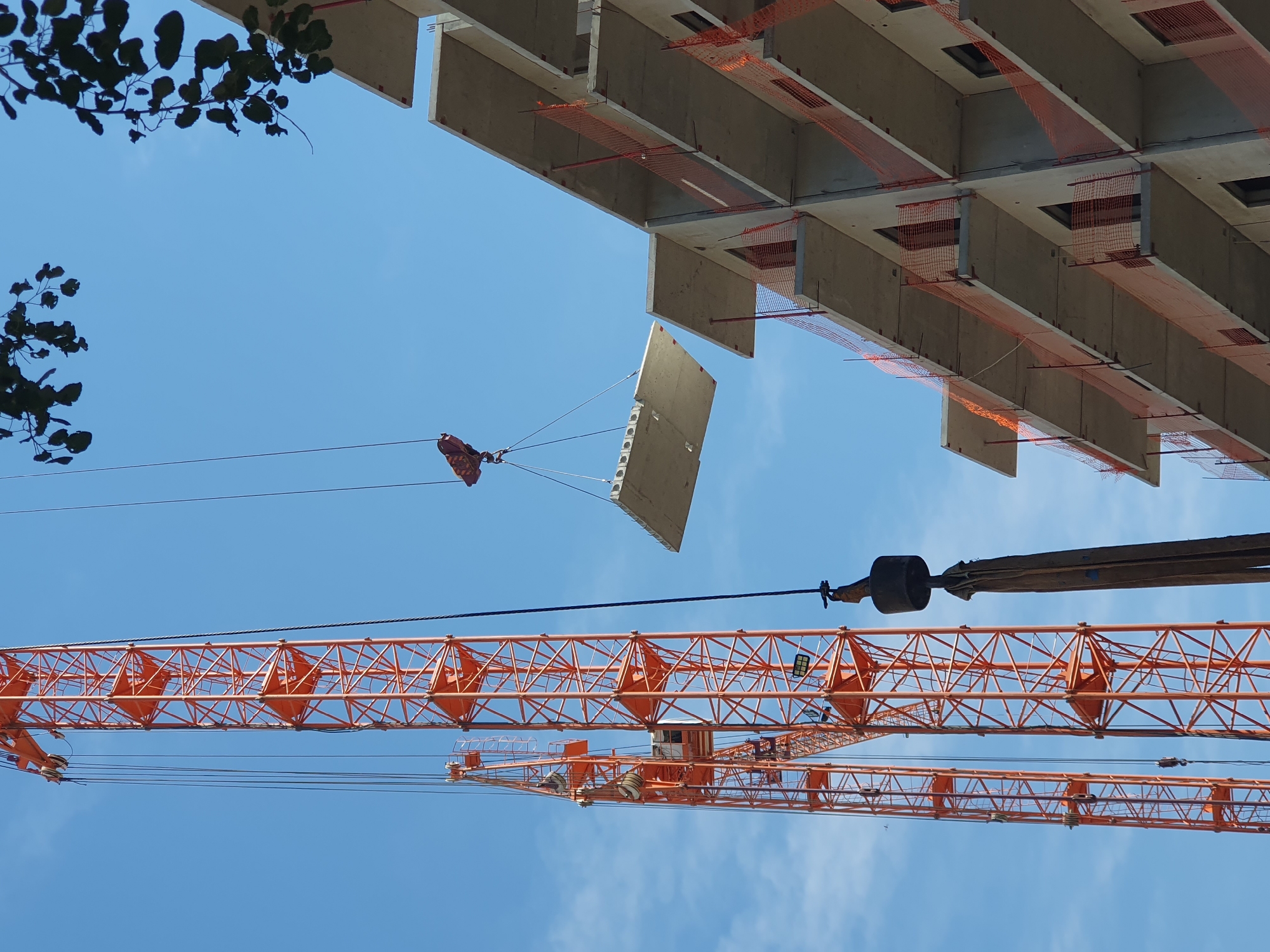 An outdoor construction scene under a clear blue sky features two orange construction cranes. The foreground on the right shows a multi-story concrete building under construction, with unfinished balconies protected by orange safety netting. A large, flat concrete slab is suspended in the air by one of the cranes, being transported to the building. The second crane, larger and taller, is visible to the left, its lattice structure extending upwards. The lower part of the scene shows the heavy lifting equipment of the crane, including a cylindrical counterweight. The image is taken from a low angle, emphasizing the scale of the construction project.