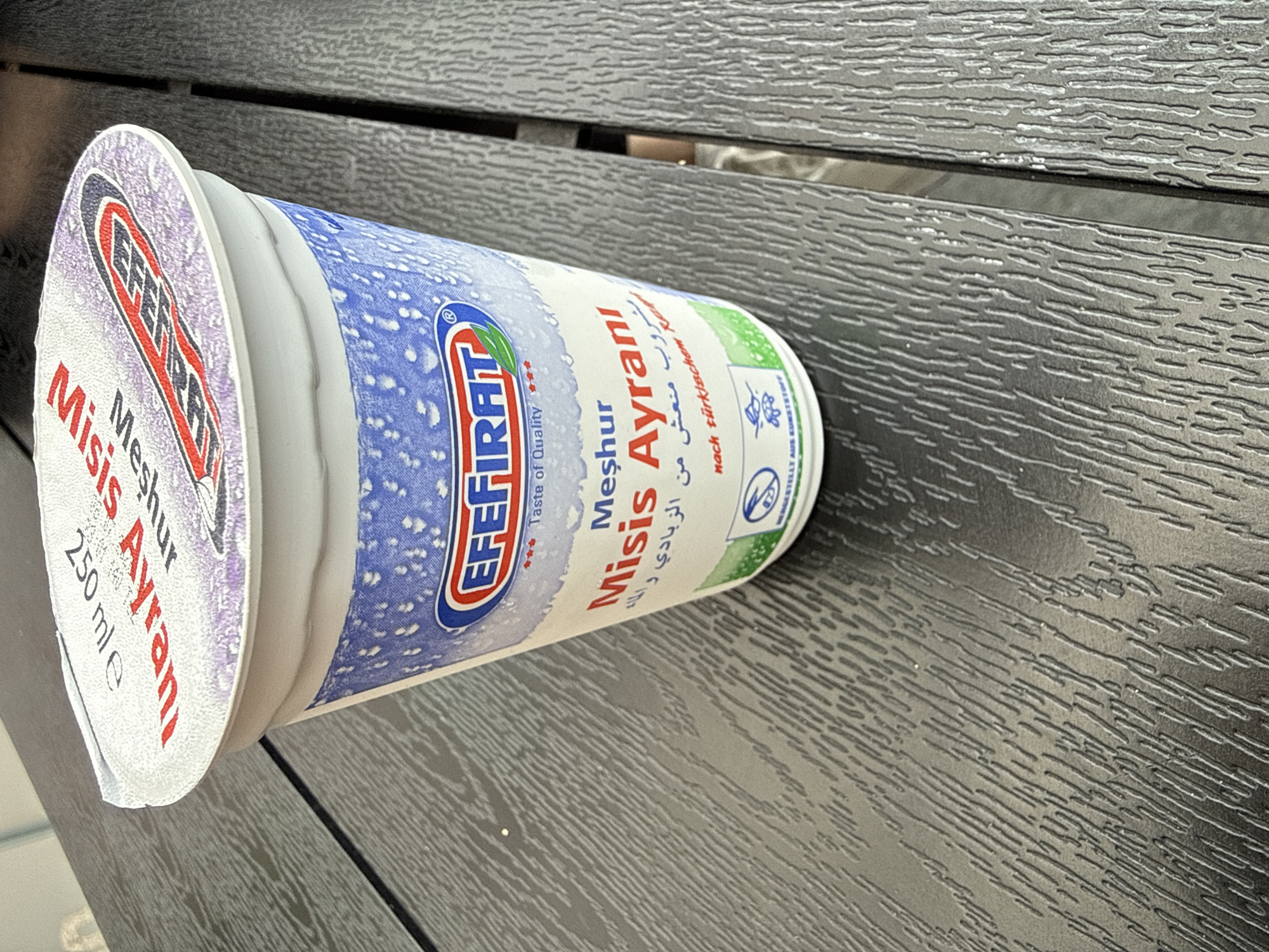 A close-up, slightly elevated shot shows a white yogurt cup with a red and white patterned lid. The lid displays text that reads "250 ml e" and "Misis Ayranı". The cup itself has a blue and white speckled design around the top, with a prominent circular logo that says "EFEFIRAT" in red, white, and blue with a green leaf. Below the logo, text reads "Taste of Quality". Further down the cup, larger text in red states "Meşhur Ayranı" followed by some Arabic script and then "nach türkischem Re". The cup is placed on a dark, textured surface, likely a table, with a visible grain pattern. The lighting suggests an outdoor or well-lit indoor environment. The image does not contain any identifiable people, specific landmarks or unique features that would pinpoint a location in the city of Großenkneten, Germany.