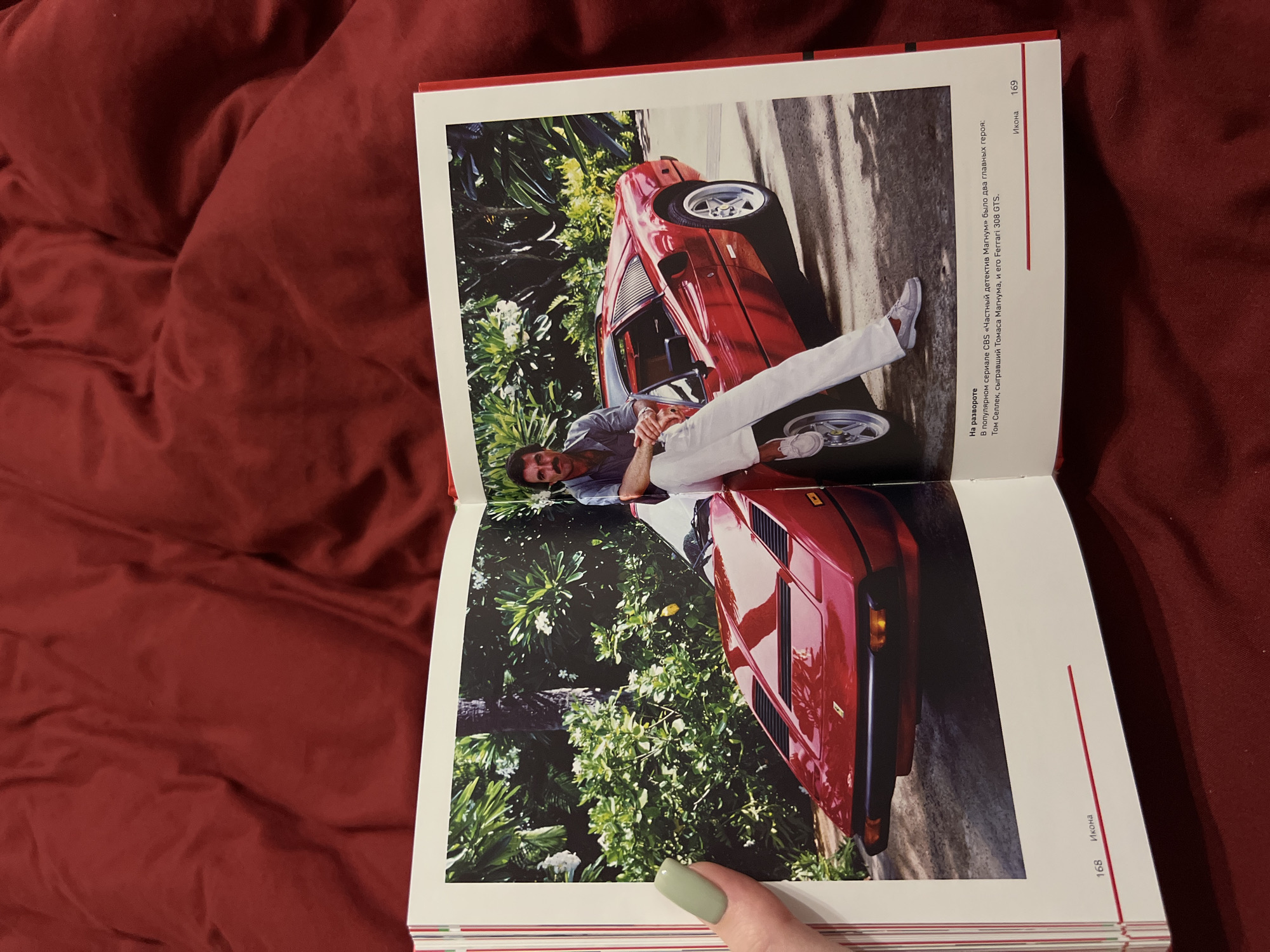 The image is a photograph of an open book resting on a textured, dark red fabric. The book displays a full-page color photograph of a man with a mustache, wearing a grey short-sleeved shirt, white pants, and light-colored loafers, sitting on a bright red Ferrari 308 GTS. The car is parked outdoors in front of lush green foliage and palm trees. Text is visible at the bottom of the page, with the number "168" on the left and "169" on the right. Below the photograph, in smaller text, there is Russian writing that translates to "On the spread. In the popular CBS series 'Magnum, P.I.' there were two main characters: Tom Selleck, who played Thomas Magnum, and his Ferrari 308 GTS." No specific location in the city of Дачное, Russia, is depicted or mentioned in the image.