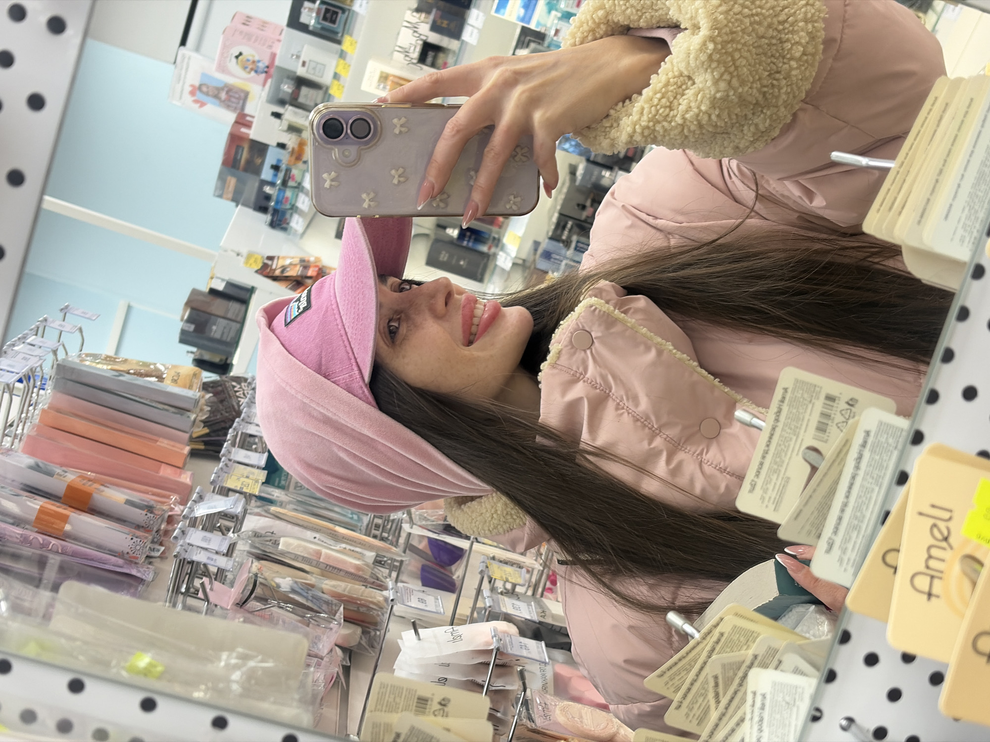 A female individual is depicted in a mirror selfie taken inside a retail store. The woman, positioned center-right, is seen from the chest up, smiling broadly with her head turned to her left. She has long, dark brown hair, fair skin with visible freckles, and light pink lipstick. She wears a light pink puffer jacket featuring a cream-colored faux shearling lining or collar, a pink velvet beanie, and a pink baseball cap with a dark emblem, worn under the beanie. Her right hand, featuring manicured nails with French tips, holds a light purple smartphone with a decorative white bow pattern on its case, which is being used to capture the mirror reflection.
The background, visible through the mirror's reflection, reveals the interior of a beauty or cosmetics store. Various products are displayed on white shelves and hanging racks. These include makeup applicators, sponges, and brushes in clear plastic packaging, as well as numerous bottles of perfume and boxed cosmetic items. A light blue wall section is visible in the upper background. In the foreground, below the individual, several product tags and packaging displaying the brand "Ameli" and Cyrillic text such as "Аппликатор д/макияжа" (Makeup applicator) are visible in the mirror's reflection. The mirror itself has a black frame with a white polka-dot pattern. Based on the Cyrillic text on the products, the location is a beauty or cosmetics store in Saint Petersburg, Russia.