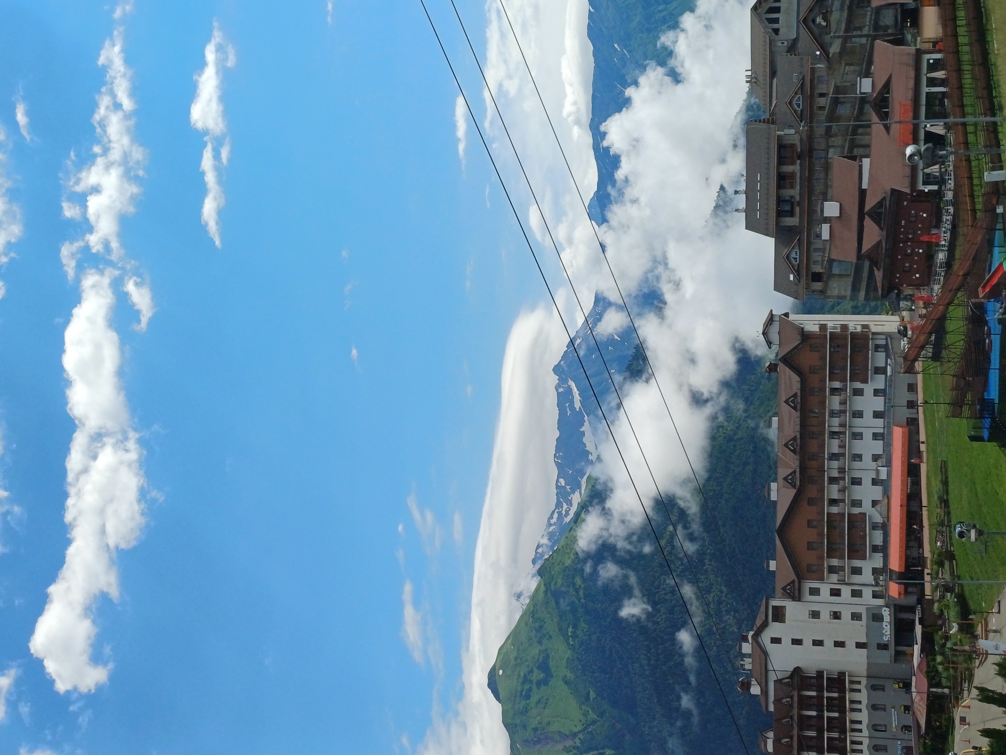The image depicts a scenic view of the mountain resort town of Estosadok, Russia, on a clear day with scattered clouds. The foreground features several multi-story buildings with brown and white facades, characteristic of mountain lodges or hotels. These structures are built on a gentle slope covered with green grass. Several cable car wires are visible stretching across the frame, suggesting a ski resort or transportation system.
In the background, lush green mountains rise dramatically, with some peaks partially obscured by white clouds that cling to their slopes. The higher mountain ridges show patches of snow, even in what appears to be a warmer season. The sky is a vibrant blue, with fluffy white clouds adding texture to the upper portion of the image.
The buildings exhibit architectural details such as balconies, dark-tiled roofs, and numerous windows. Some of the buildings have orange awnings over ground-level entrances or patios. There are some areas of outdoor seating and recreational features visible, such as a small playground with a red slide. The overall impression is one of a well-developed tourist destination nestled within a stunning natural landscape.