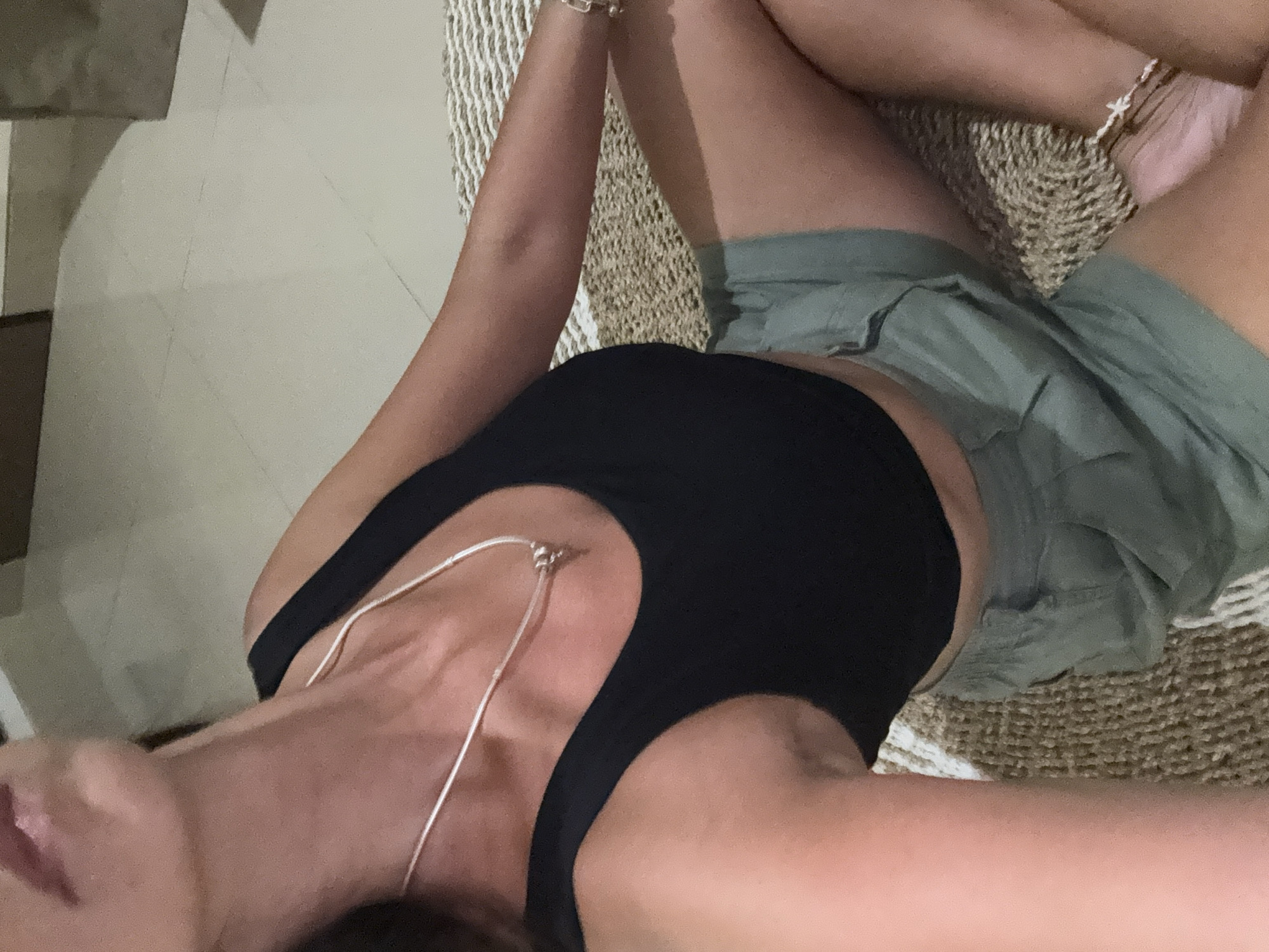 The image shows a person lying on their back, viewed from a slightly elevated perspective. The subject is wearing a black sleeveless top and olive green shorts. A delicate silver chain necklace is visible on their chest. On their right wrist, multiple bracelets are stacked, and a beaded anklet with small white charms, possibly crosses, adorns their right ankle.
The person is resting on a textured woven mat, which features a pattern of light brown and white stripes. Parts of a light-colored tiled floor with subtle diagonal lines are visible to the left and beneath the mat. The lighting is soft and diffused, suggesting an indoor setting, a shaded area, or possibly evening light. The person's head is partially visible on the left, with their mouth slightly open, conveying a relaxed and serene expression. Their arms are bent and resting, and their legs are also bent and crossed at the ankles, contributing to the overall relaxed posture. No specific activity other than resting is depicted. There is no visible text. The scene suggests a calm environment, fitting the context of a tropical location like Kuta Selatan, Indonesia.