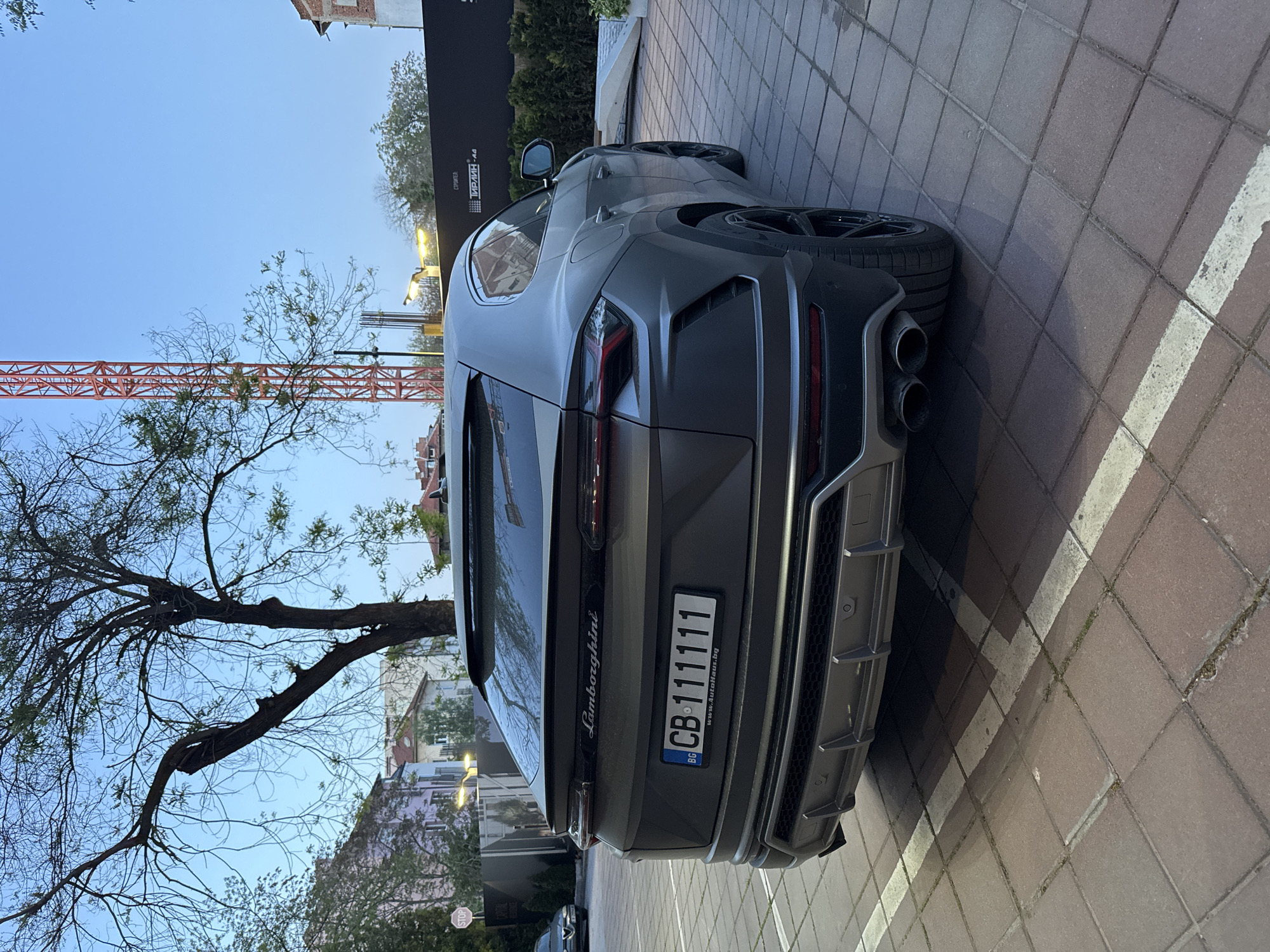 A dark gray Lamborghini Urus SUV is parked on a paved surface in an urban setting during twilight hours in Plovdiv, Bulgaria. The vehicle is seen from its rear-right side, prominently displaying its distinctive design elements such as the LED taillights, quad exhaust tips, and a sculpted rear diffuser. A Bulgarian license plate, "CB 111111", is affixed to the tailgate, along with "BG" and "www.autohaus.bg". The word "Lamborghini" is visible above the license plate.
The car rests on reddish-brown interlocking pavers that feature white painted lines, suggesting a parking area or roadside. In the background, a large, bare-branched tree occupies the left side, while greener trees and foliage are scattered throughout. An orange construction crane is partially visible against the light blue sky, indicating ongoing development. To the right of the crane, a large black sign displays Cyrillic text, including "СТРОИТЕЛ:" (Builder:) and "ИРИН .АД", likely identifying a construction company. A lit streetlight emits a warm glow in the mid-ground, and a small "STOP" sign is partially visible near the bottom right. The overall scene suggests a quiet, unoccupied urban environment at dusk or dawn.